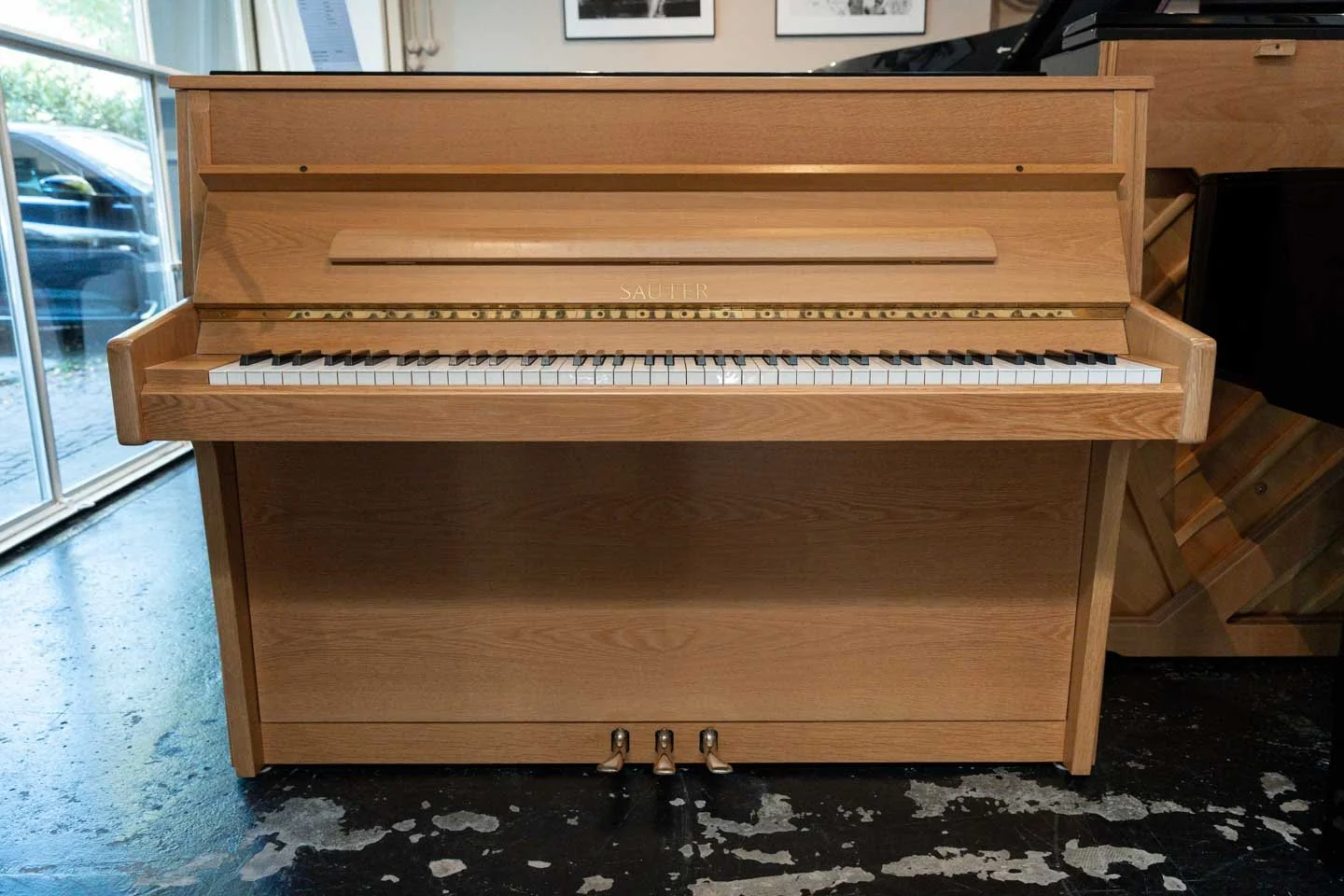 Ein Holzklavier in einem Raum, mit einer Pedalerie am Boden und einem Fenster im Hintergrund.