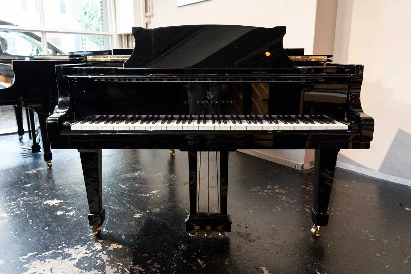 Ein schwarzes Flügelklavier der Marke Steinway & Sons in einem Raum mit Holzboden und Fenstern im Hintergrund.