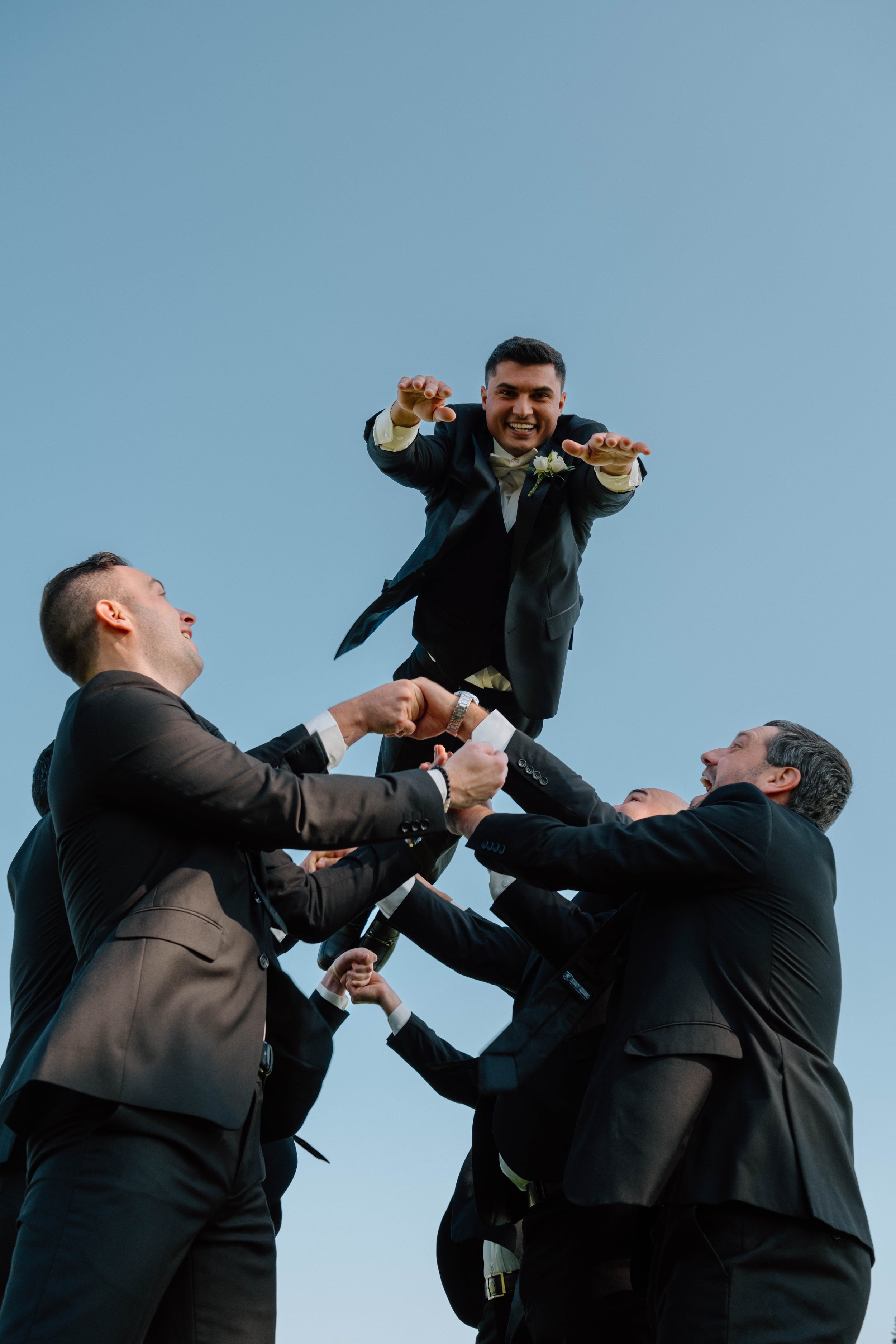 Group of men in suits lifting a man in a tuxedo into the air against a blue sky, celebrating a special occasion.