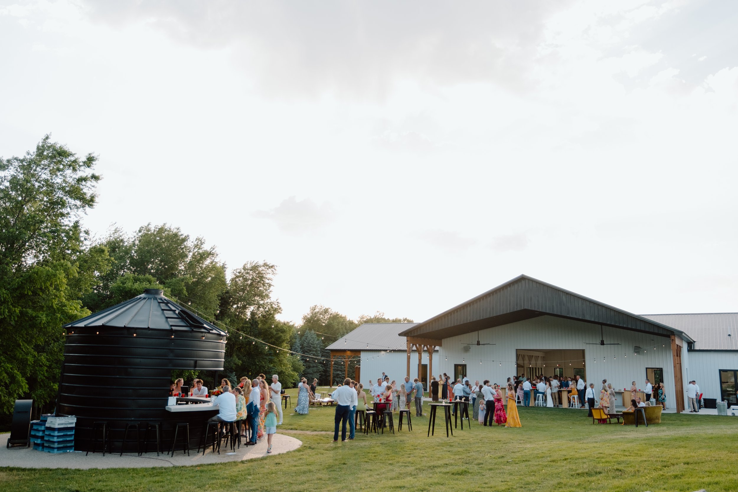People socializing outdoors at a wedding reception in Northern Michigan with a barn, trees, and a black silo, during the evening.