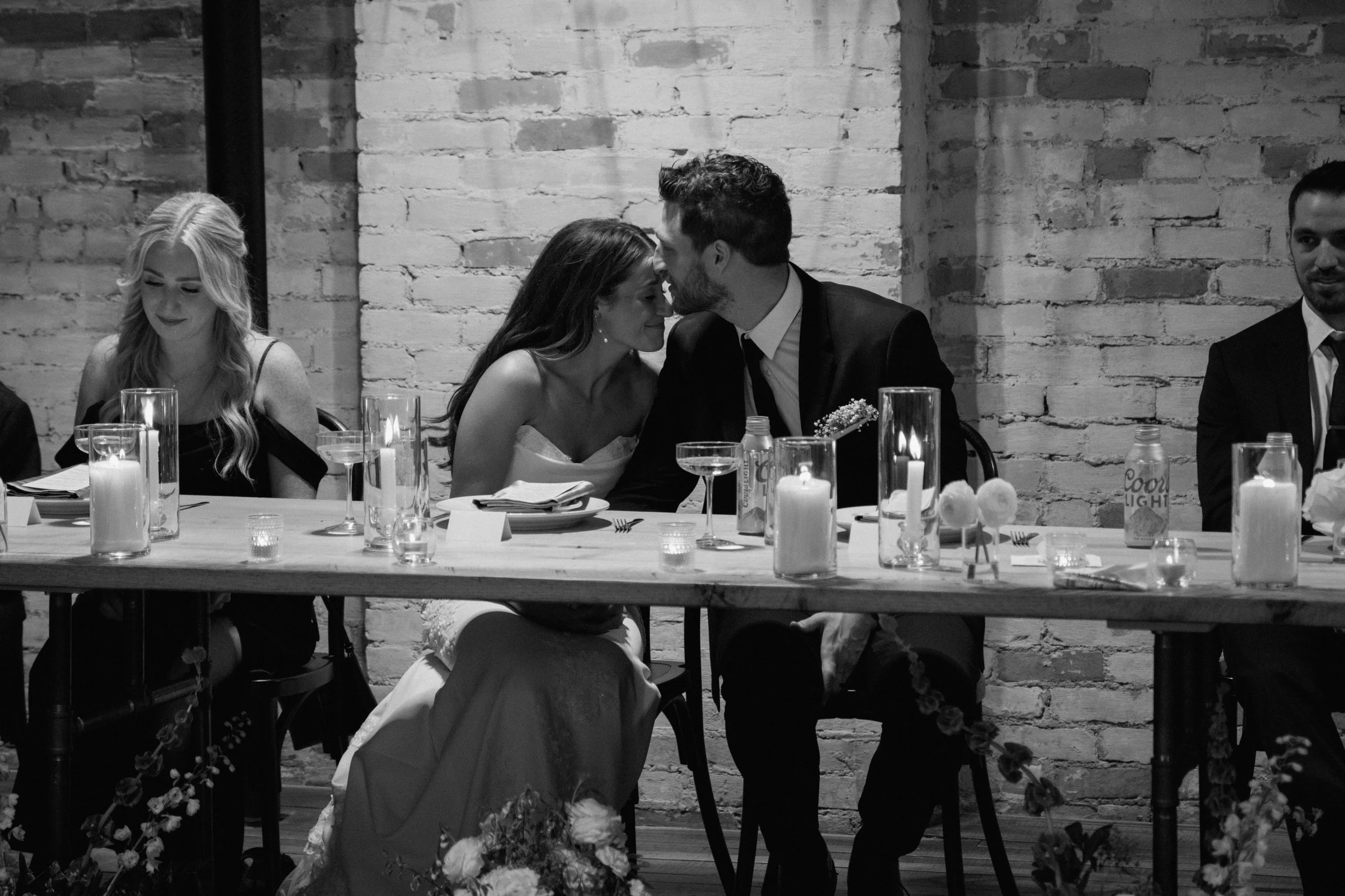 Black and white photo of a wedding reception with a bride and groom sharing a moment at the table surrounded by friends and decorated with candles and flowers.