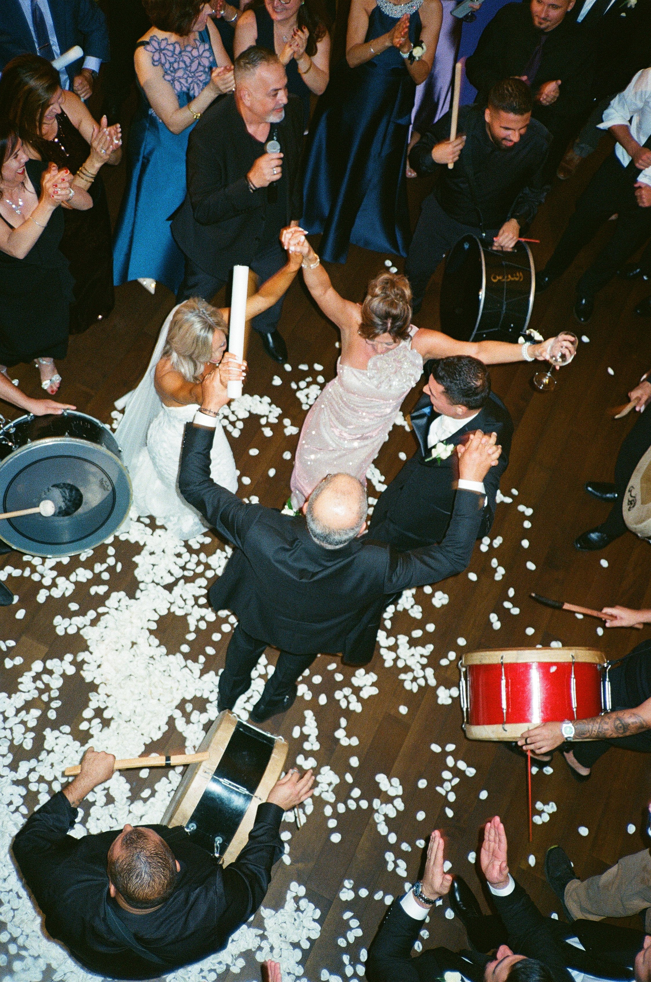 A wedding celebration with the bride and groom dancing in the center surrounded by guests, some playing drums, with flower petals on the floor.
