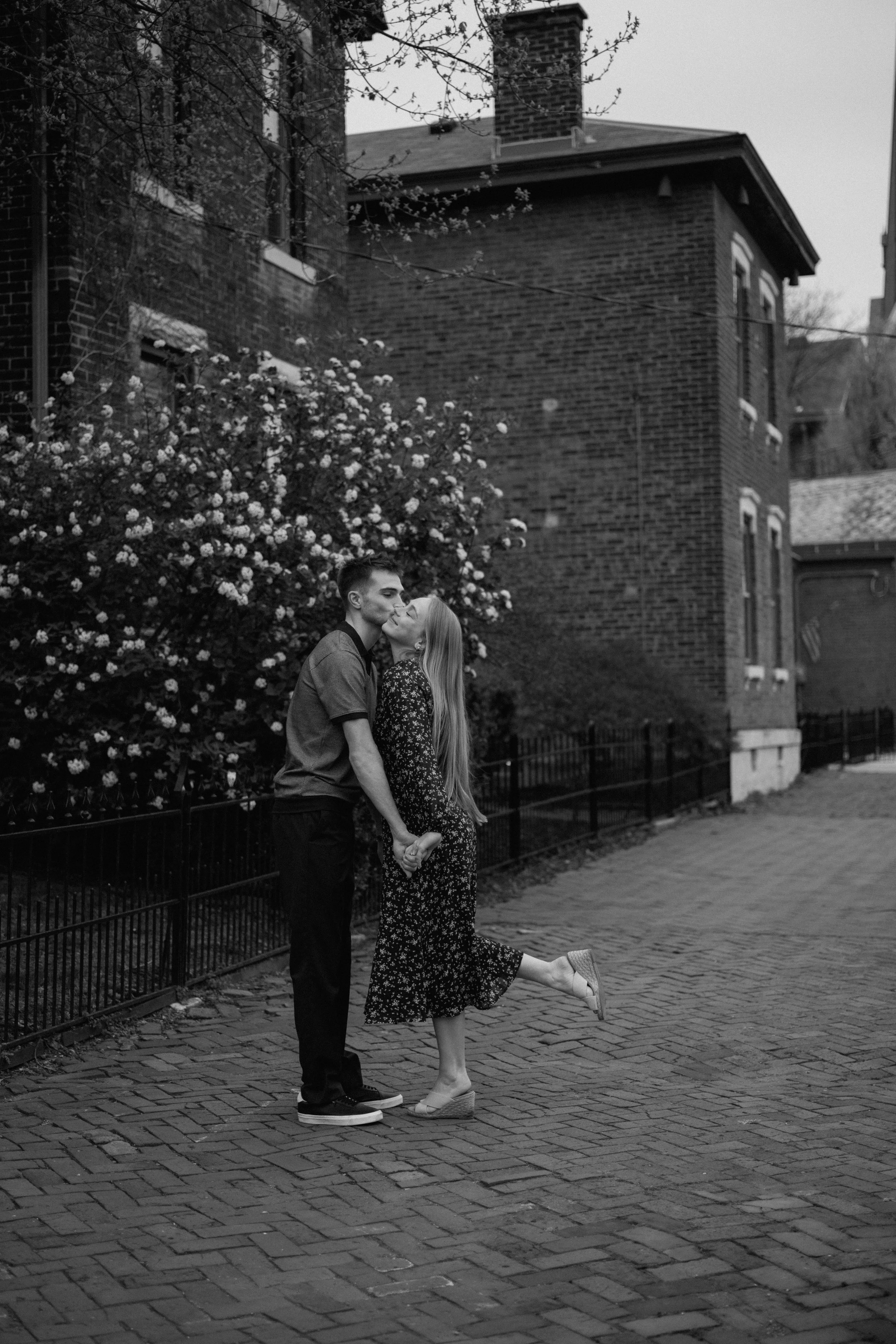 A couple kissing outdoors on a cobblestone street, holding hands, with a flowering bush and brick houses in Columbus, Ohio.