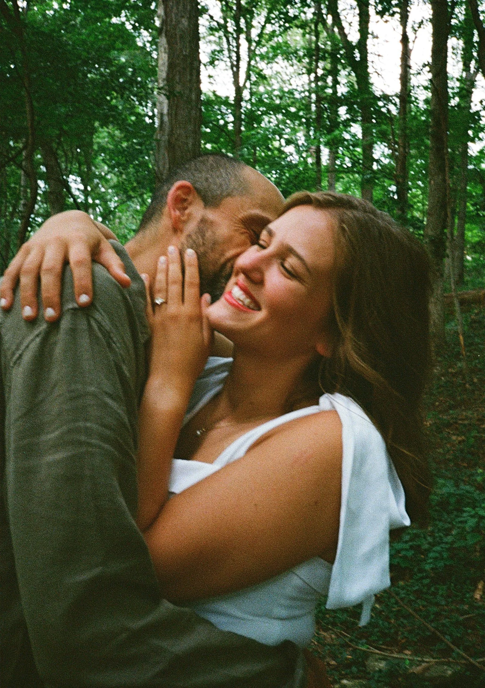 A film photo of a happy couple hugging and smiling in a forest, with trees and greenery in the background.
