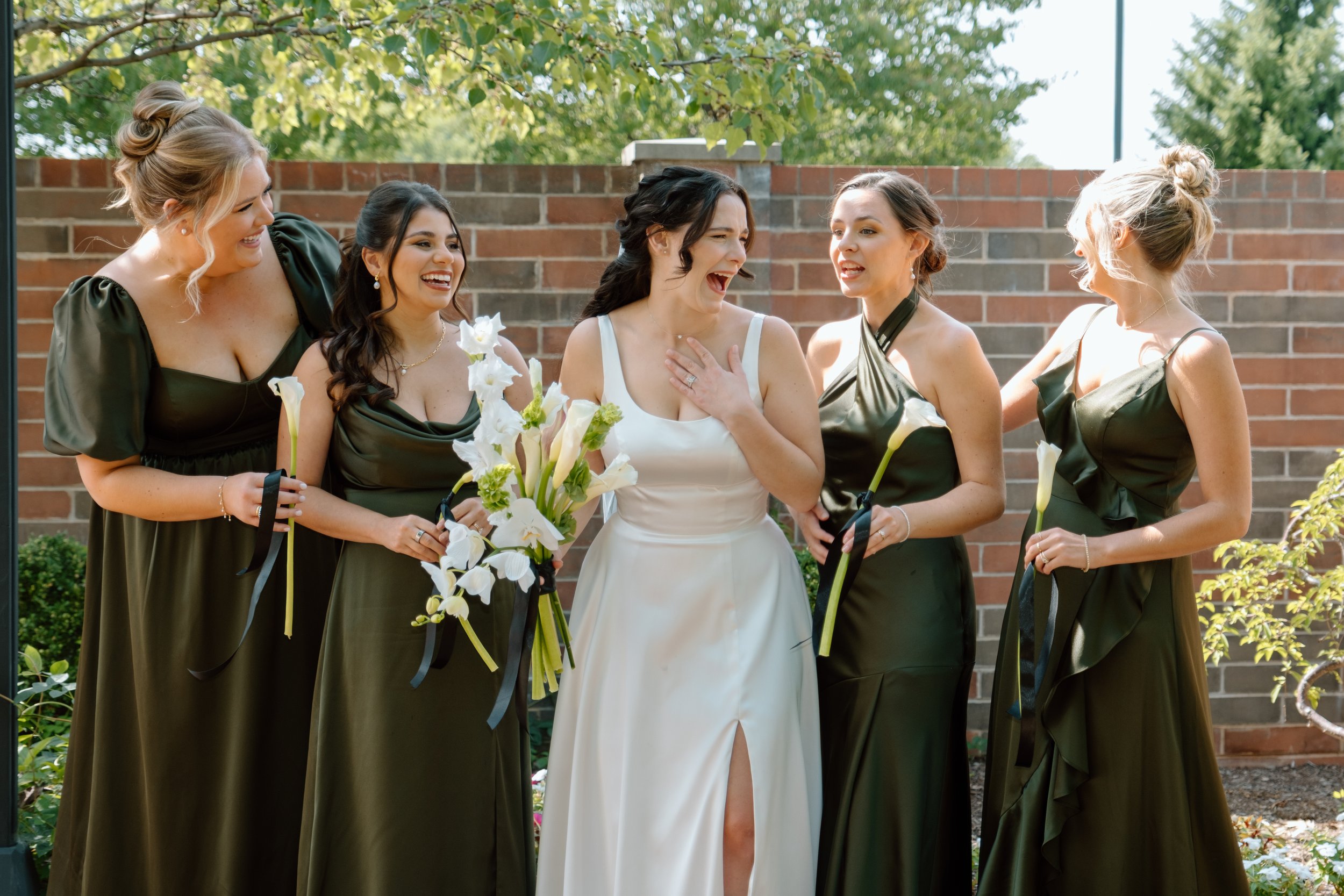 A bride in a white dress and five bridesmaids in dark green dresses laugh outdoors in front of a brick wall, holding flowers.