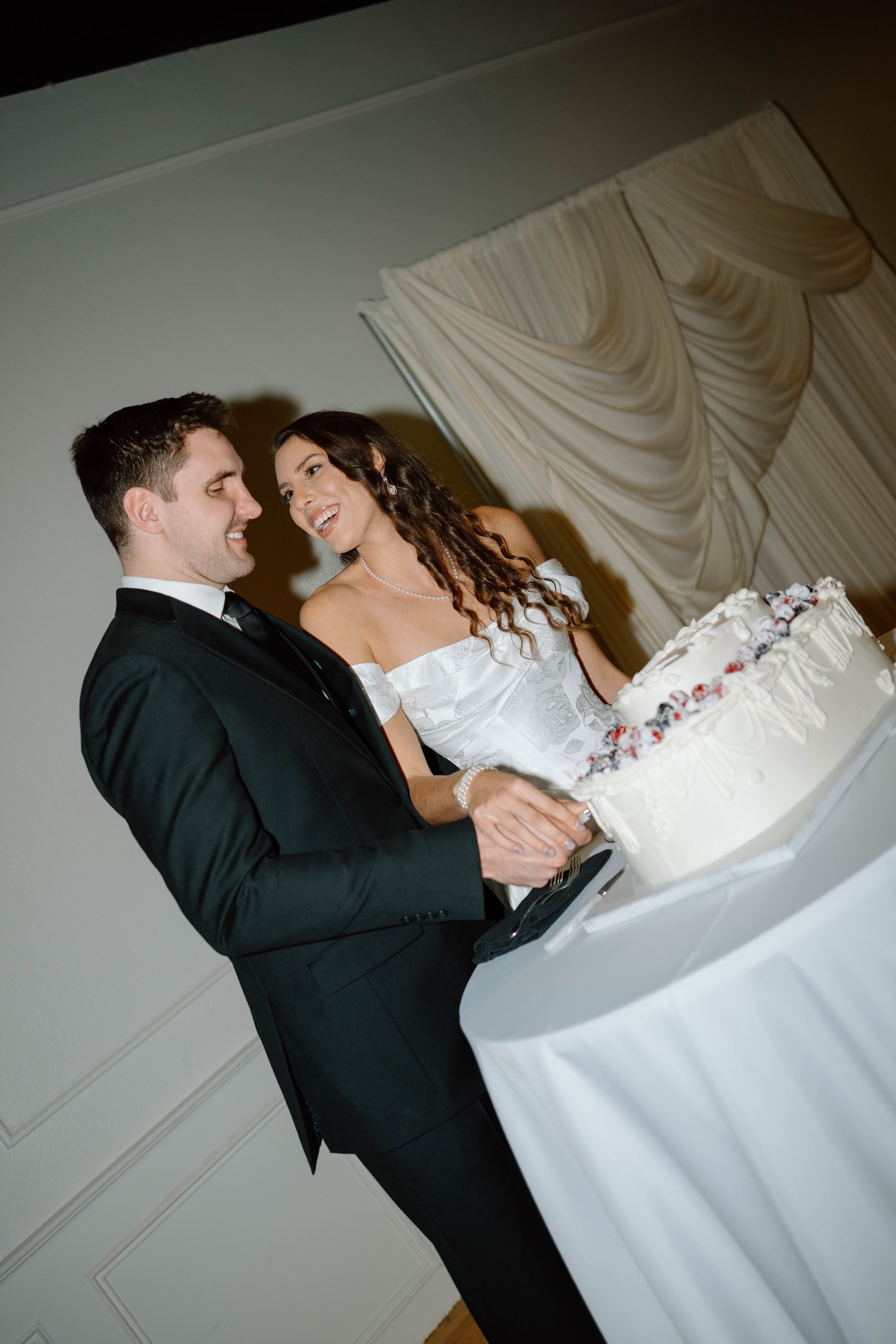 A bride and groom cutting their wedding cake, smiling at each other indoors.