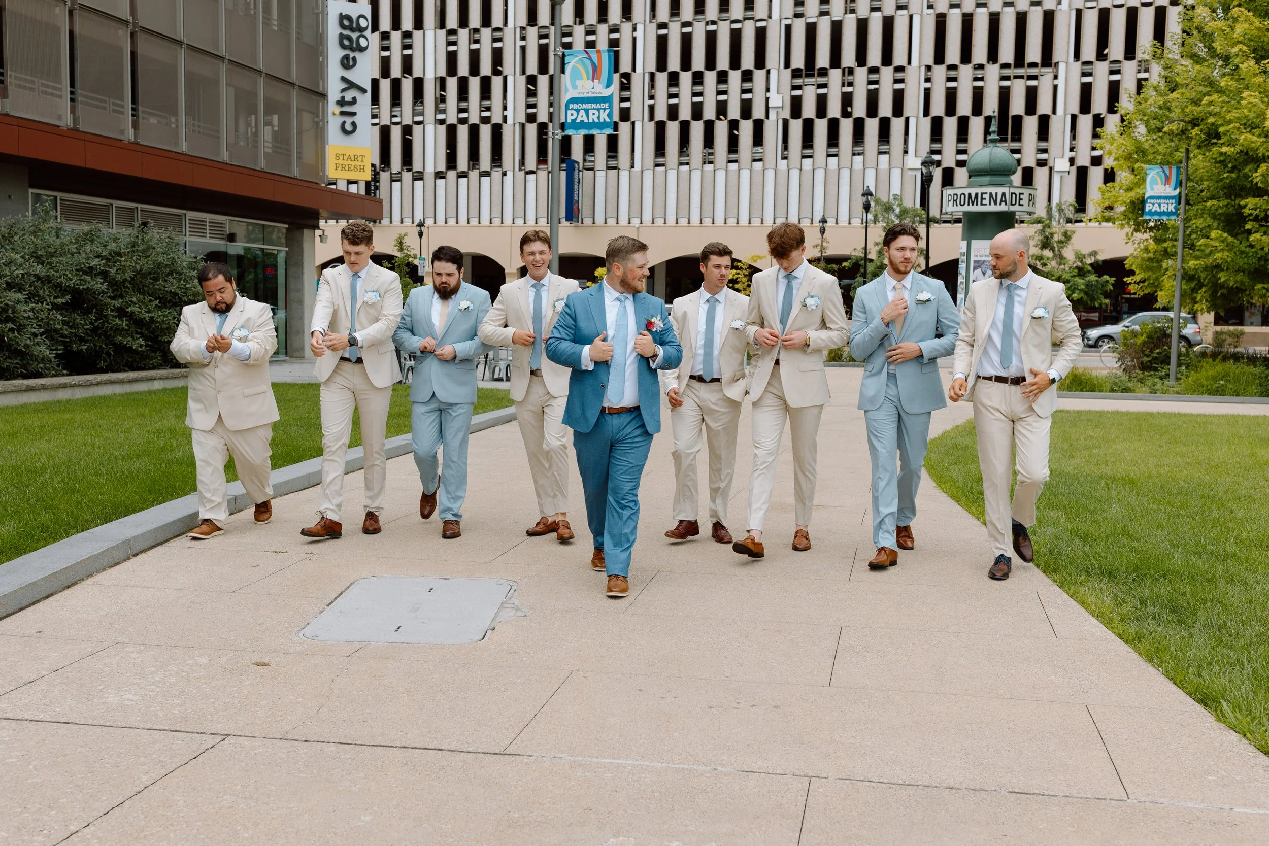 A group of men dressed in suits walking on a sidewalk in an urban area, with some adjusting their jackets or looking at their phones.