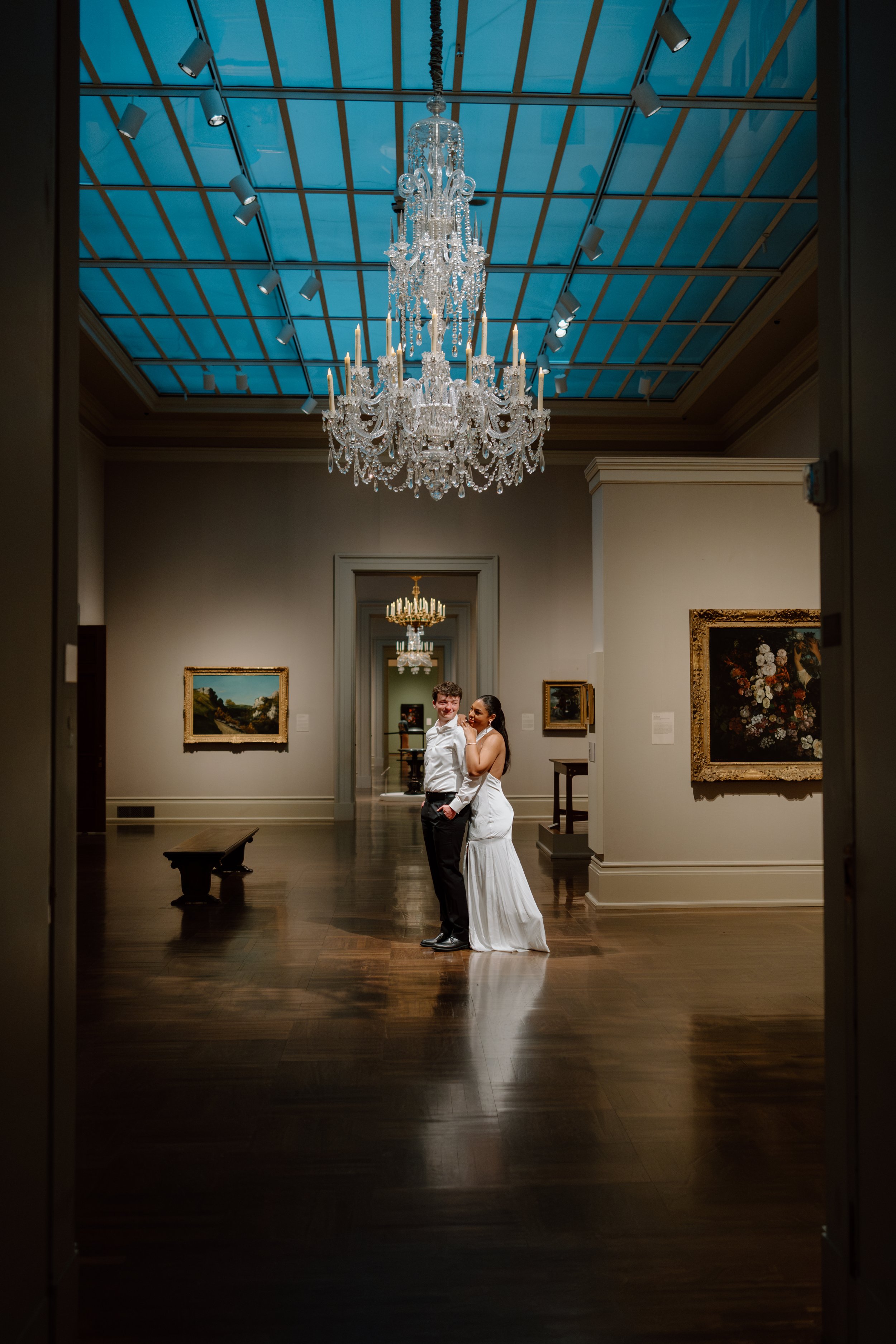 A bride and groom standing together in Toledo Museum of Art, framed by an ornate doorway, under a large crystal chandelier on a high ceiling with track lighting, surrounded by framed paintings, with a wooden bench to the left.