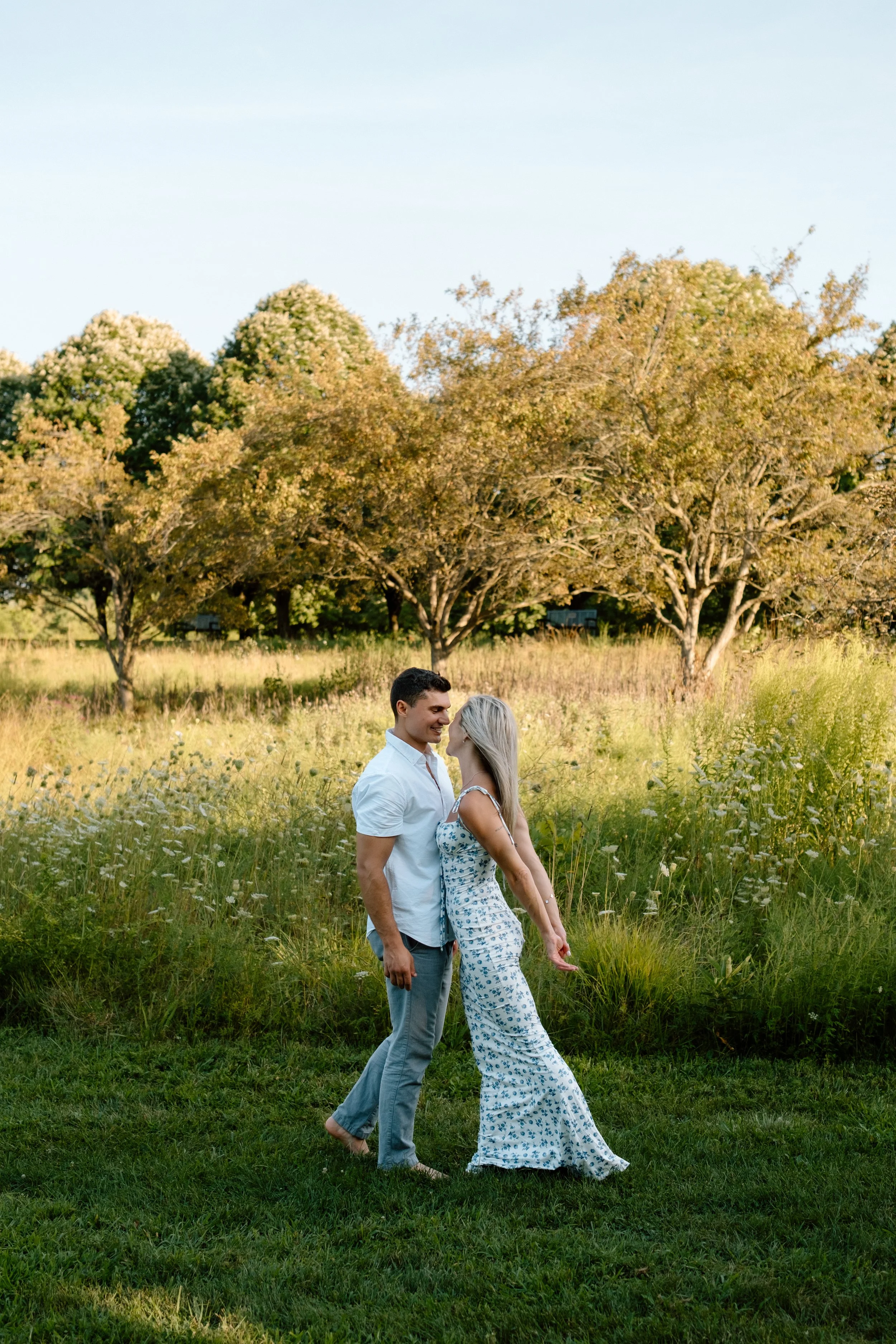 A couple standing closely in a grassy field with trees in the background during golden hour.