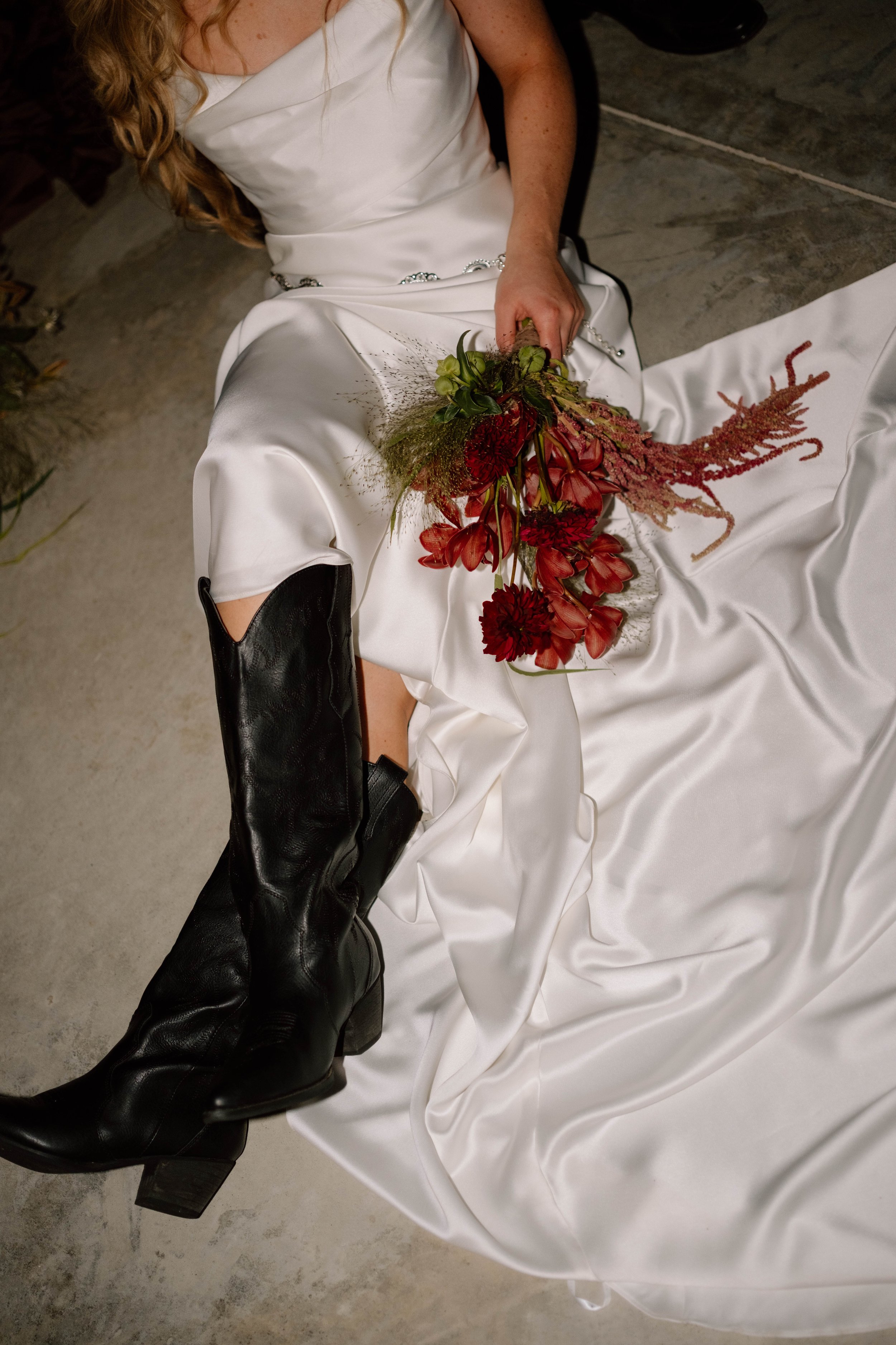 A woman in a white dress, sitting on the floor with black cowboy boots, holding a bouquet of red and green flowers, with a white satin fabric draped around her.
