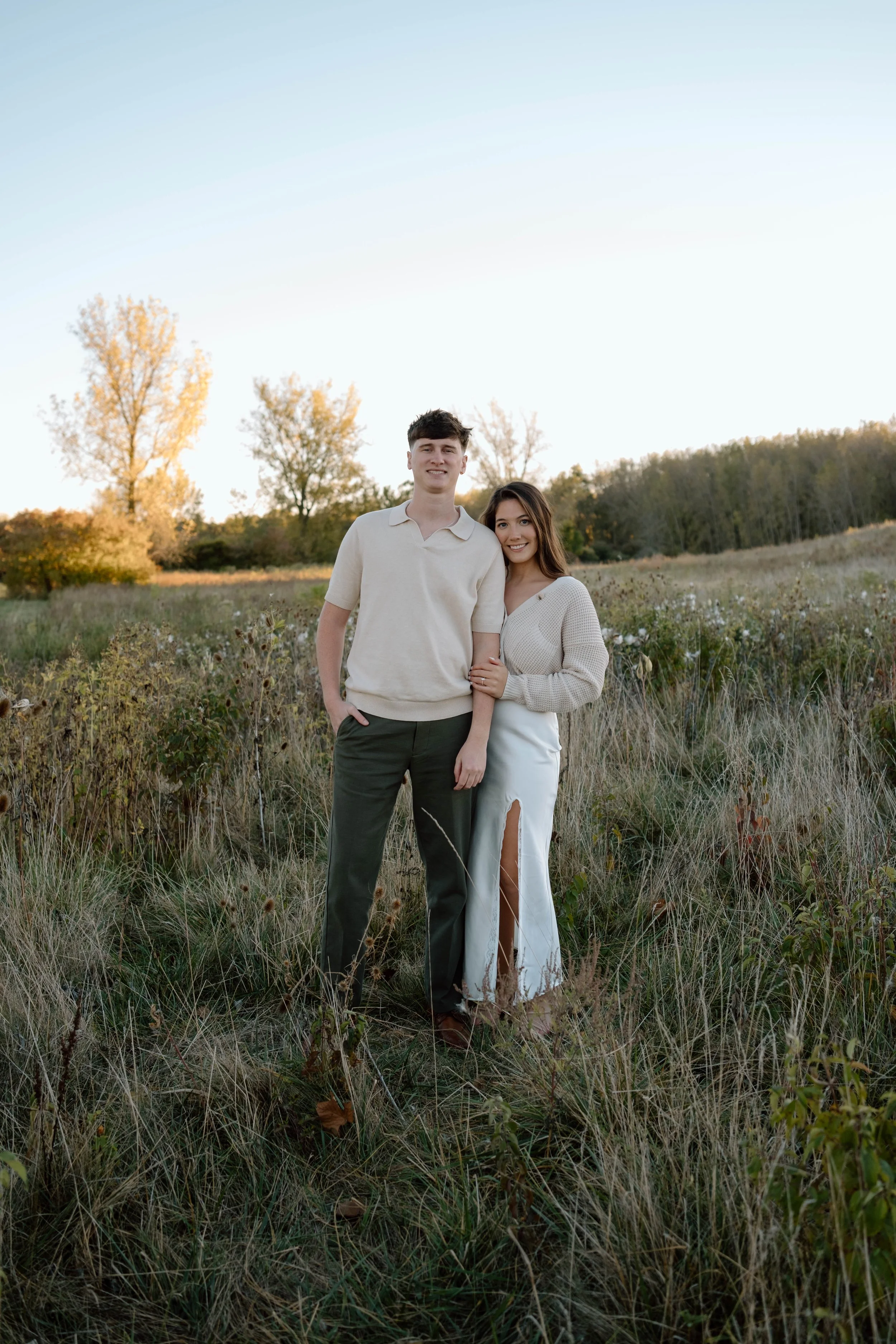 A young couple standing together in a grassy field during the daytime, smiling at the camera with trees in the background.