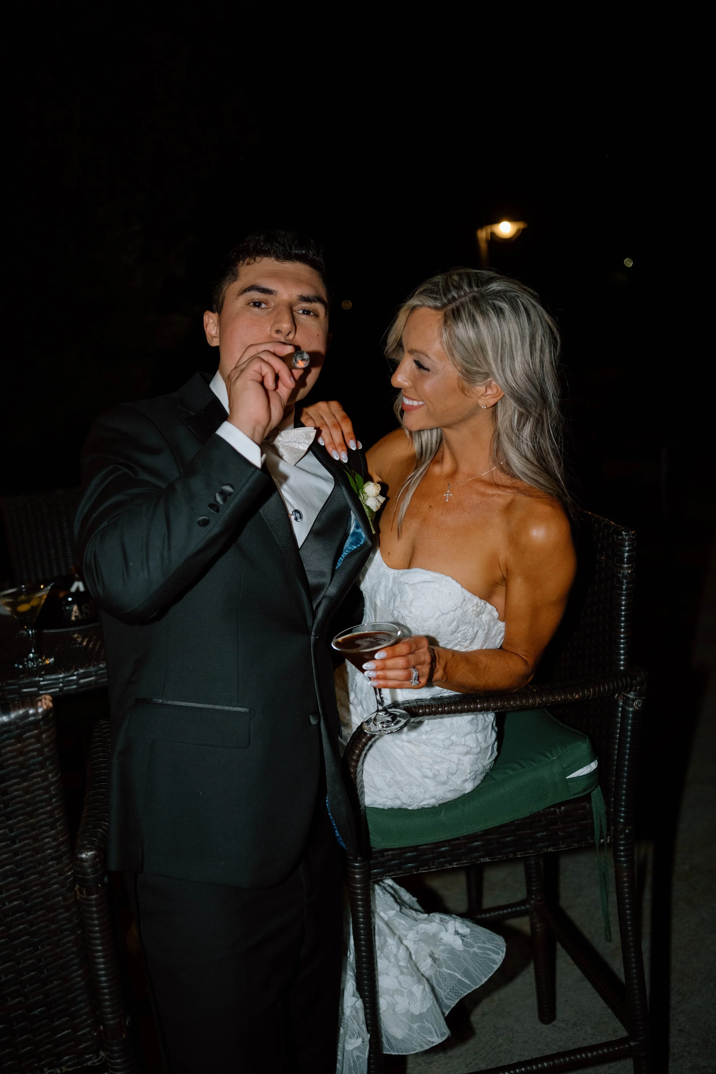 A man in a tuxedo is smoking a cigar while a woman in a white dress sits on a chair holding a cocktail glass, both at a nighttime outdoor event.