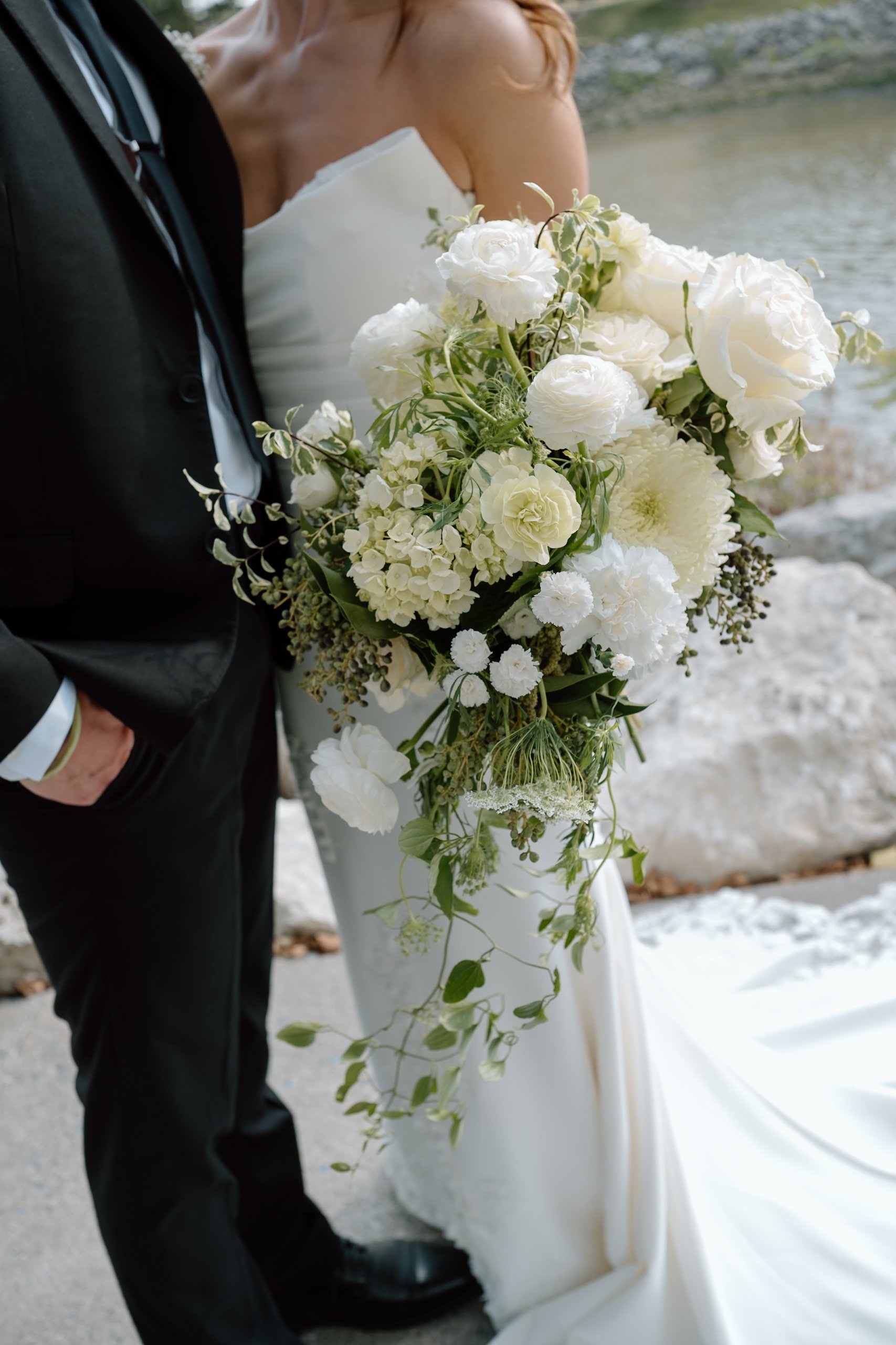 A bride holding a cascading white flower bouquet beside her groom at a waterfront.