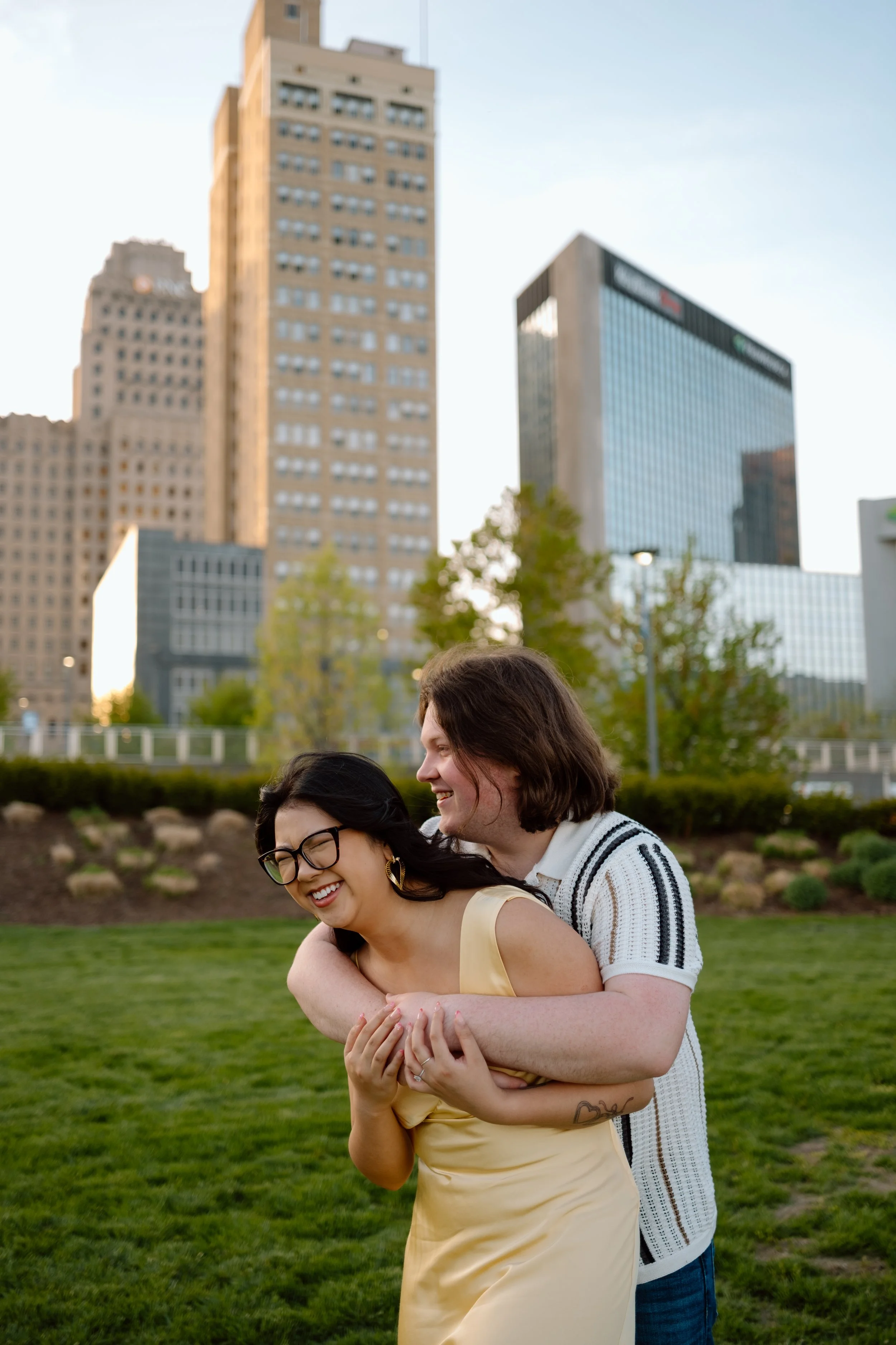 A couple laughing and hugging in a park with Toledo's skyline in the background.