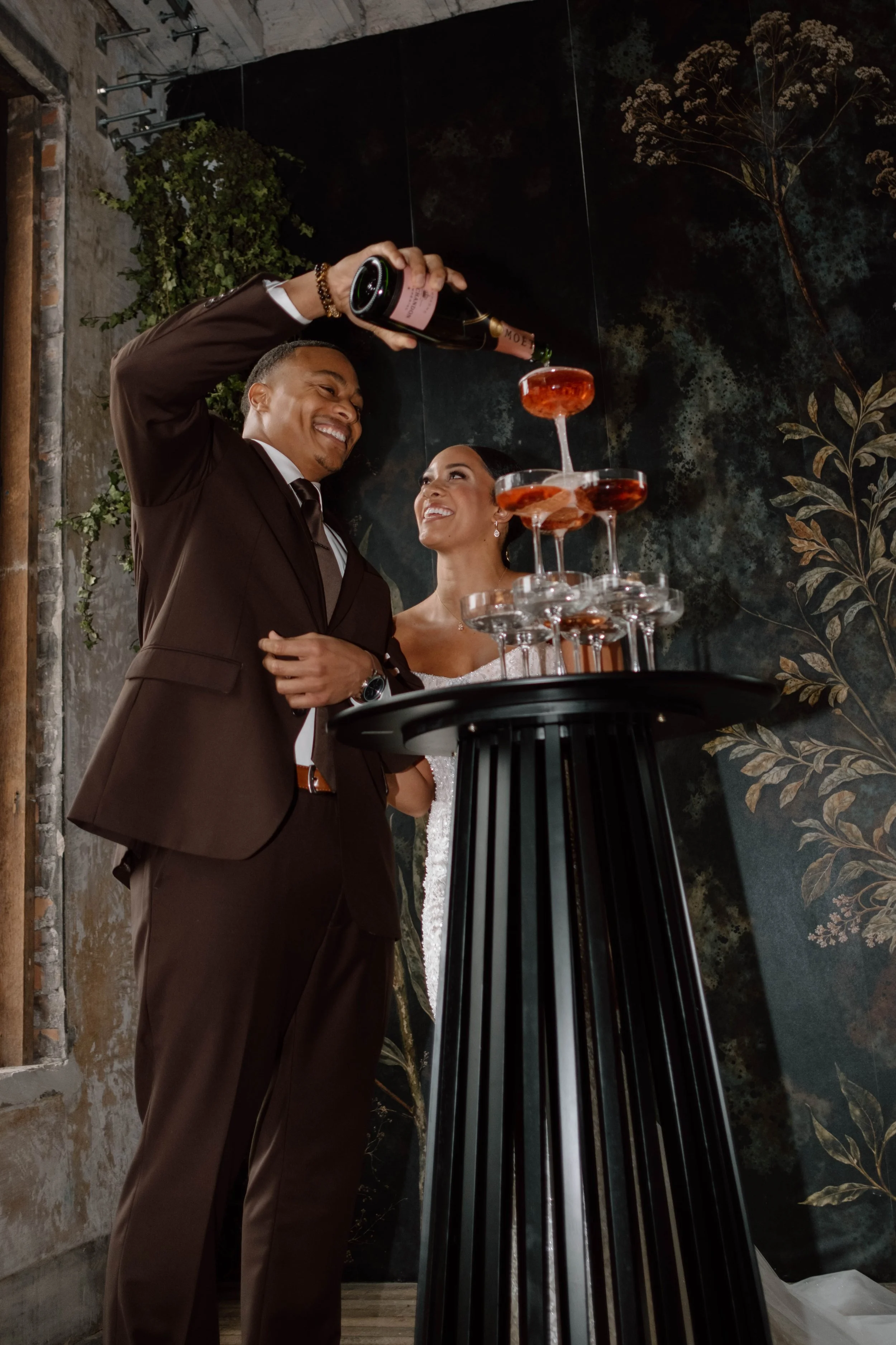 A couple in wedding attire enjoying a champagne toast at their wedding reception, with a tower of glasses filled with drinks and a dark floral wallpaper background.