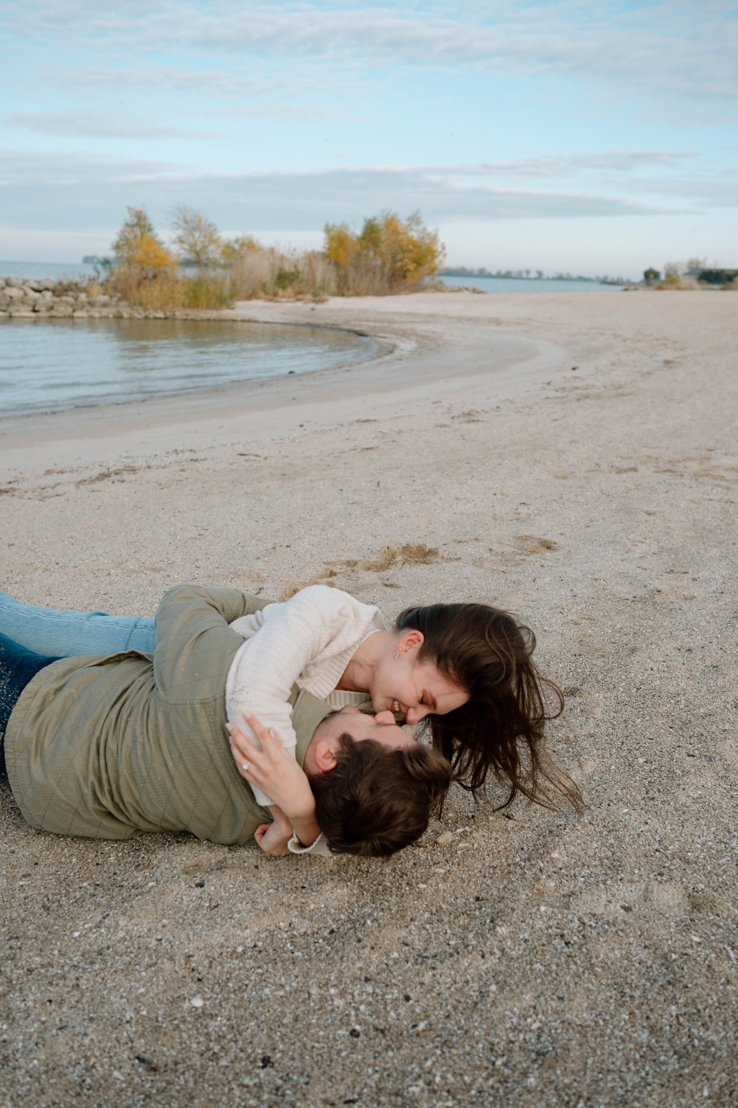A couple laying on a sandy beach, playfully engaged in a close embrace, with the woman leaning over the man, both smiling, near water and trees in the background.