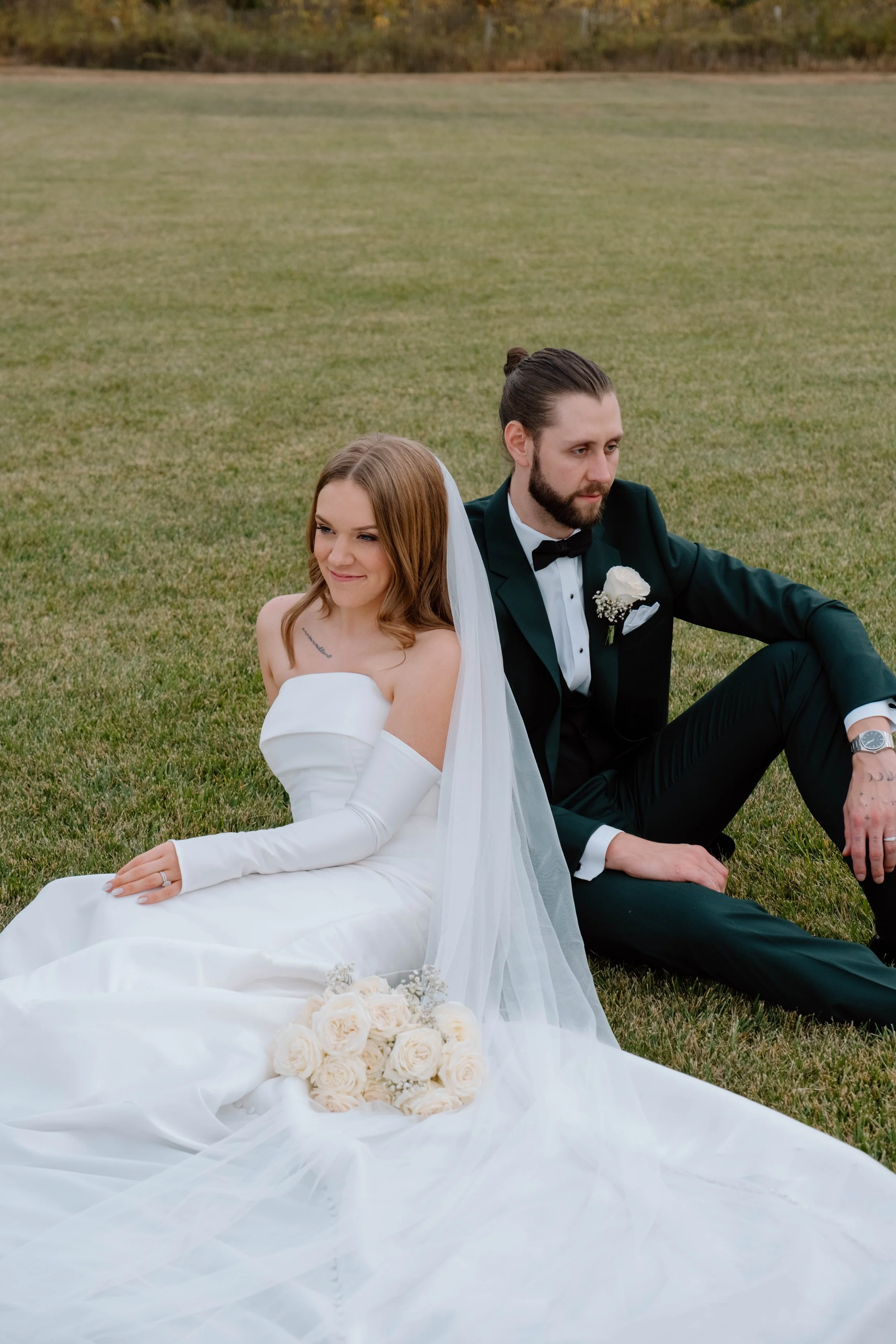 A bride and groom sitting on the grass. The bride is in a white wedding gown with long gloves, holding a bouquet of white roses. The groom is in a black tuxedo with a bow tie, sitting with one knee up, looking off to the side.