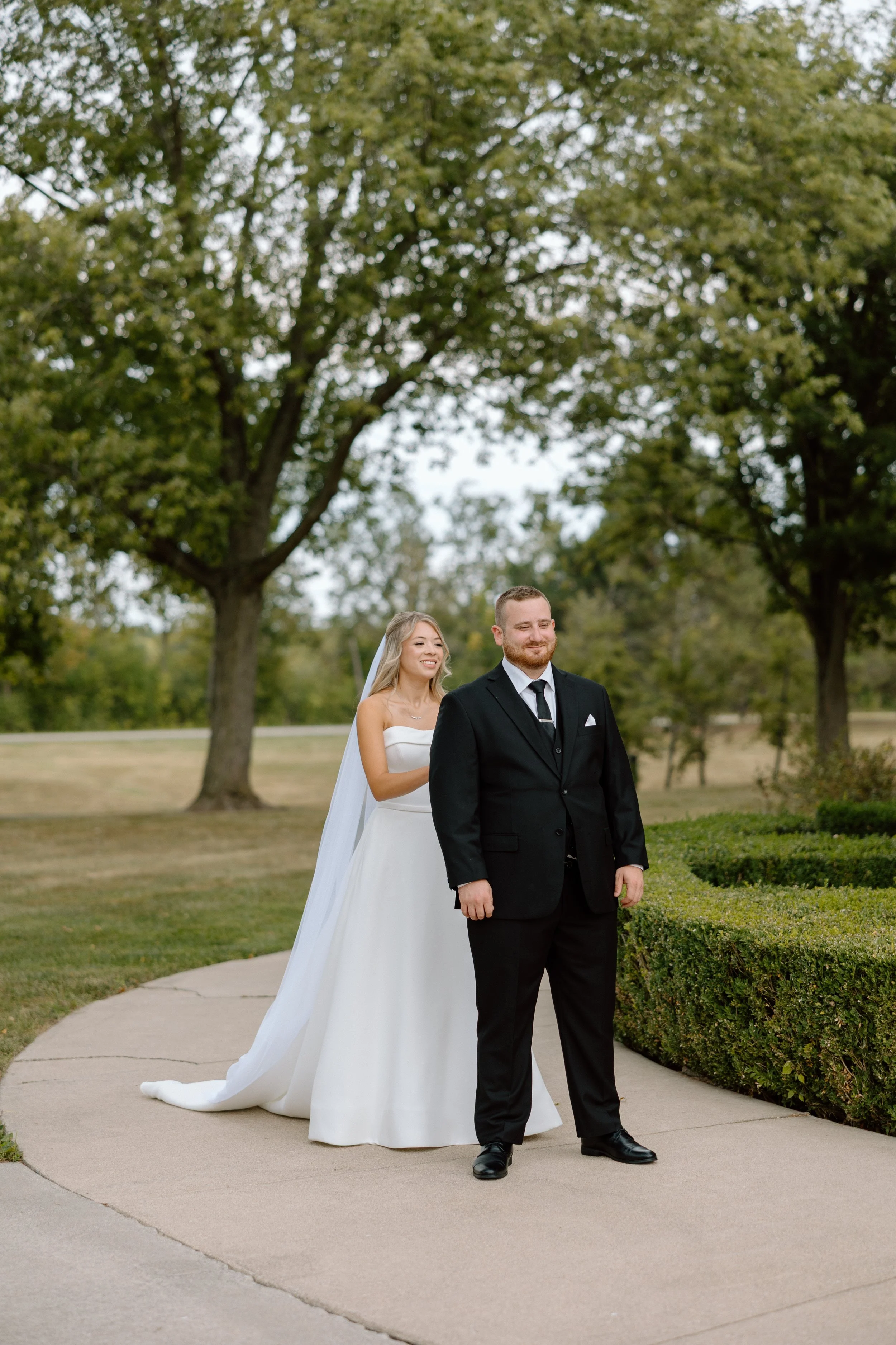 A bride and groom standing on a sidewalk outdoors during daytime, with the bride smiling and dressed in a white wedding gown with a veil, and the groom in a black suit with a white shirt and black tie, surrounded by trees and greenery.