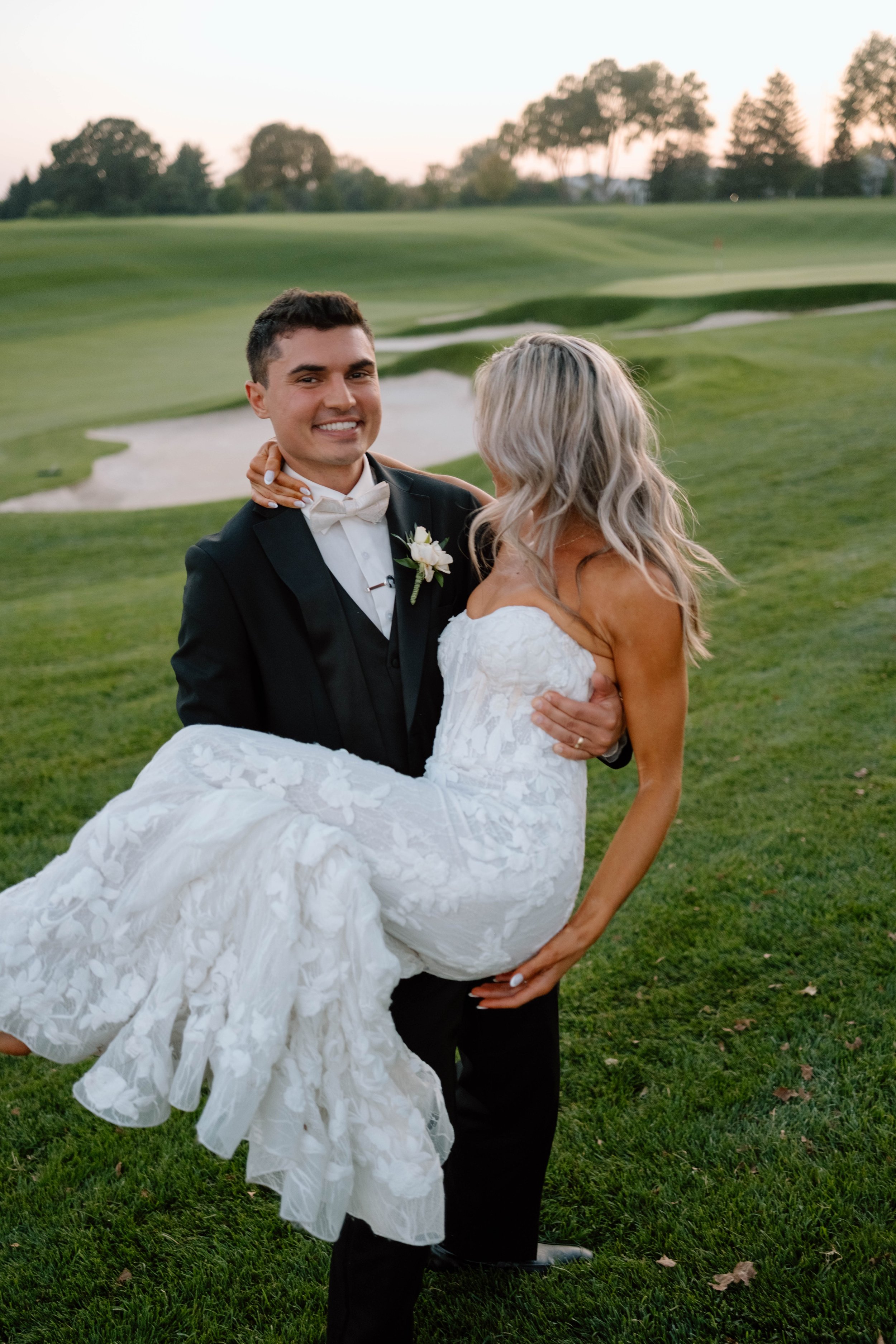 A groom in a black tuxedo holding a bride in a white wedding dress on a golf course at sunset.