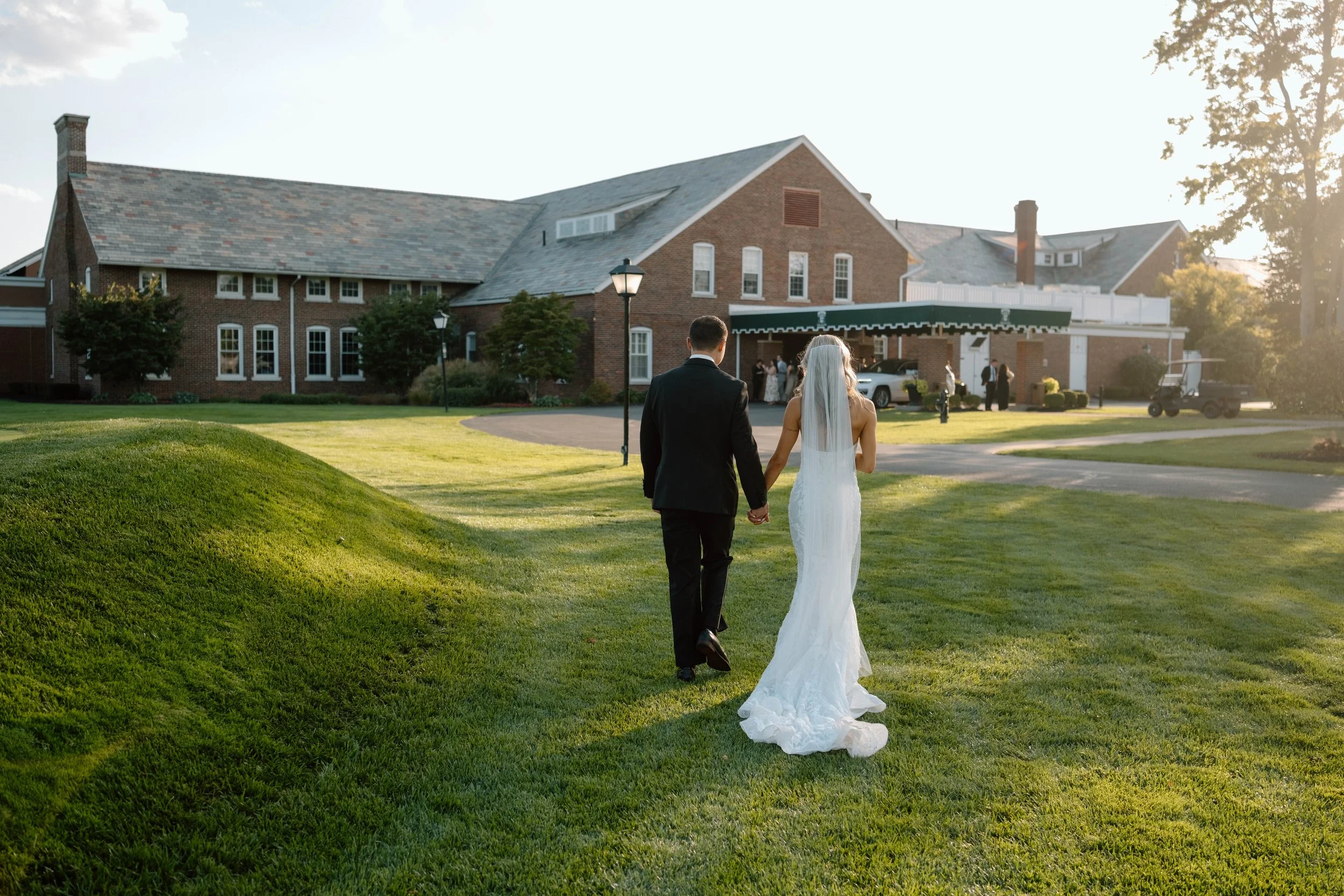 A bride and groom holding hands walk toward a large brick building, likely a wedding venue, on a sunny day with green grass and a clear sky.