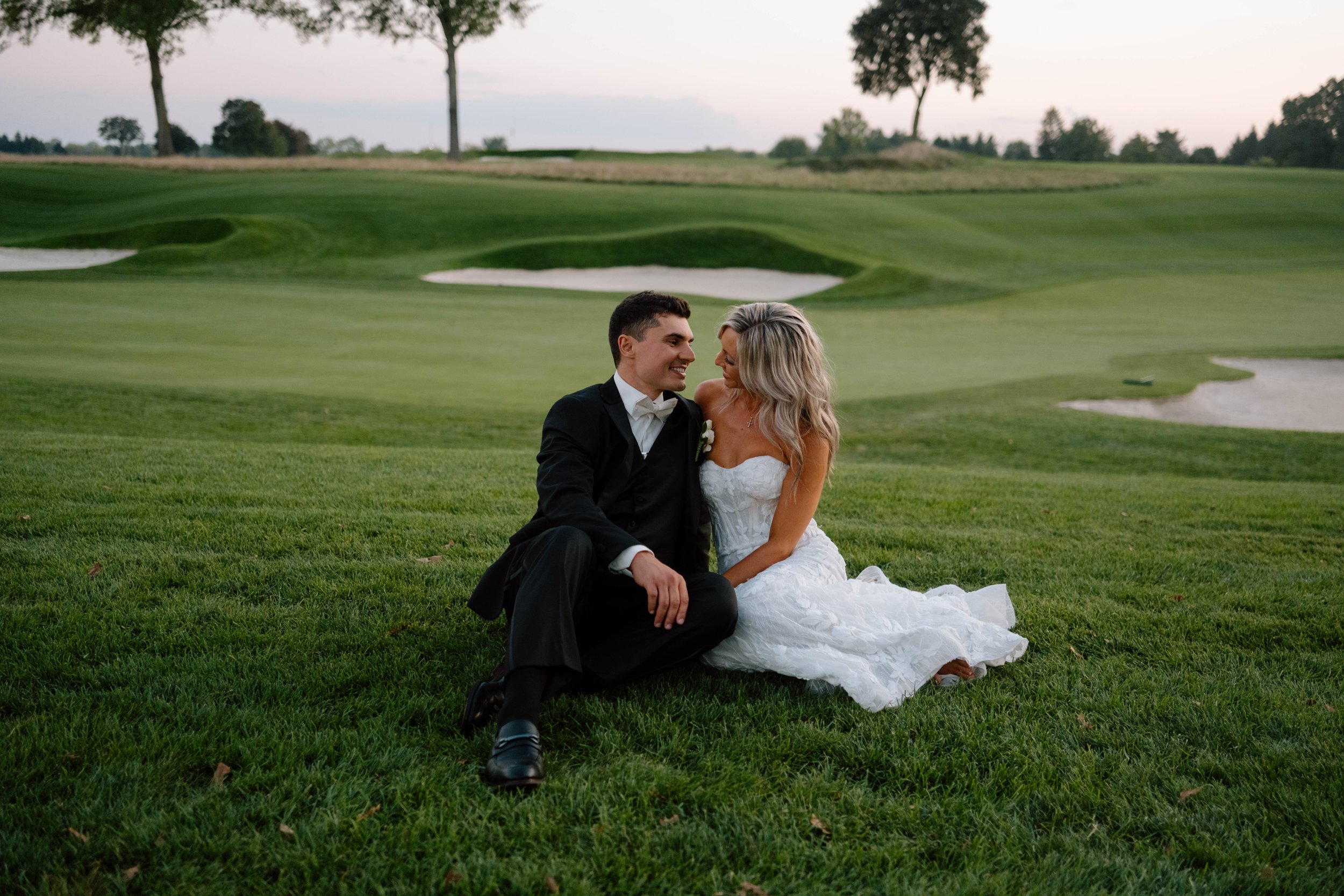 A bride and groom sitting on a golf course grass, looking at each other and smiling, with sand bunkers and trees in the background.