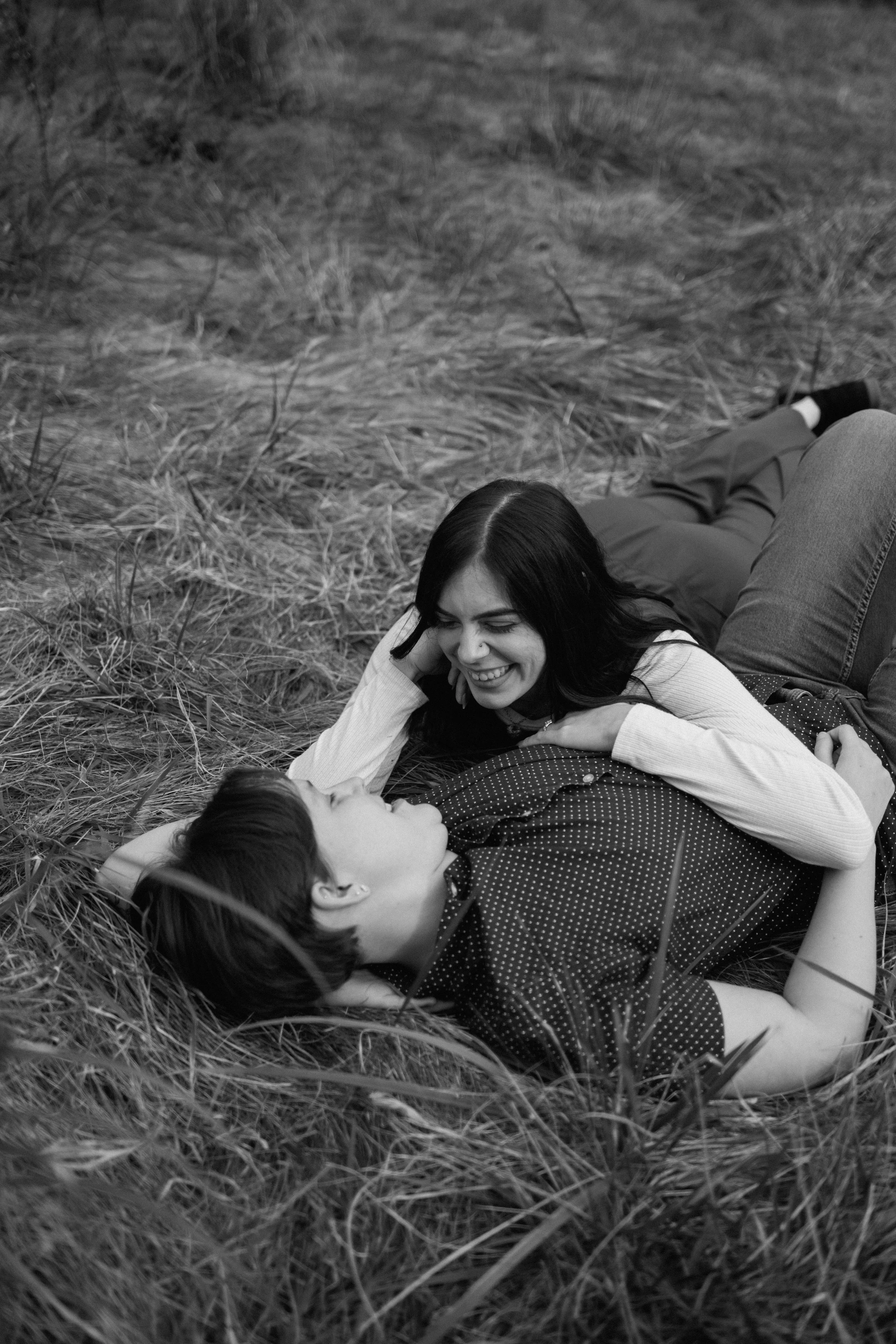 A woman and a man lying on the ground in a field of tall grass, smiling and looking at each other.