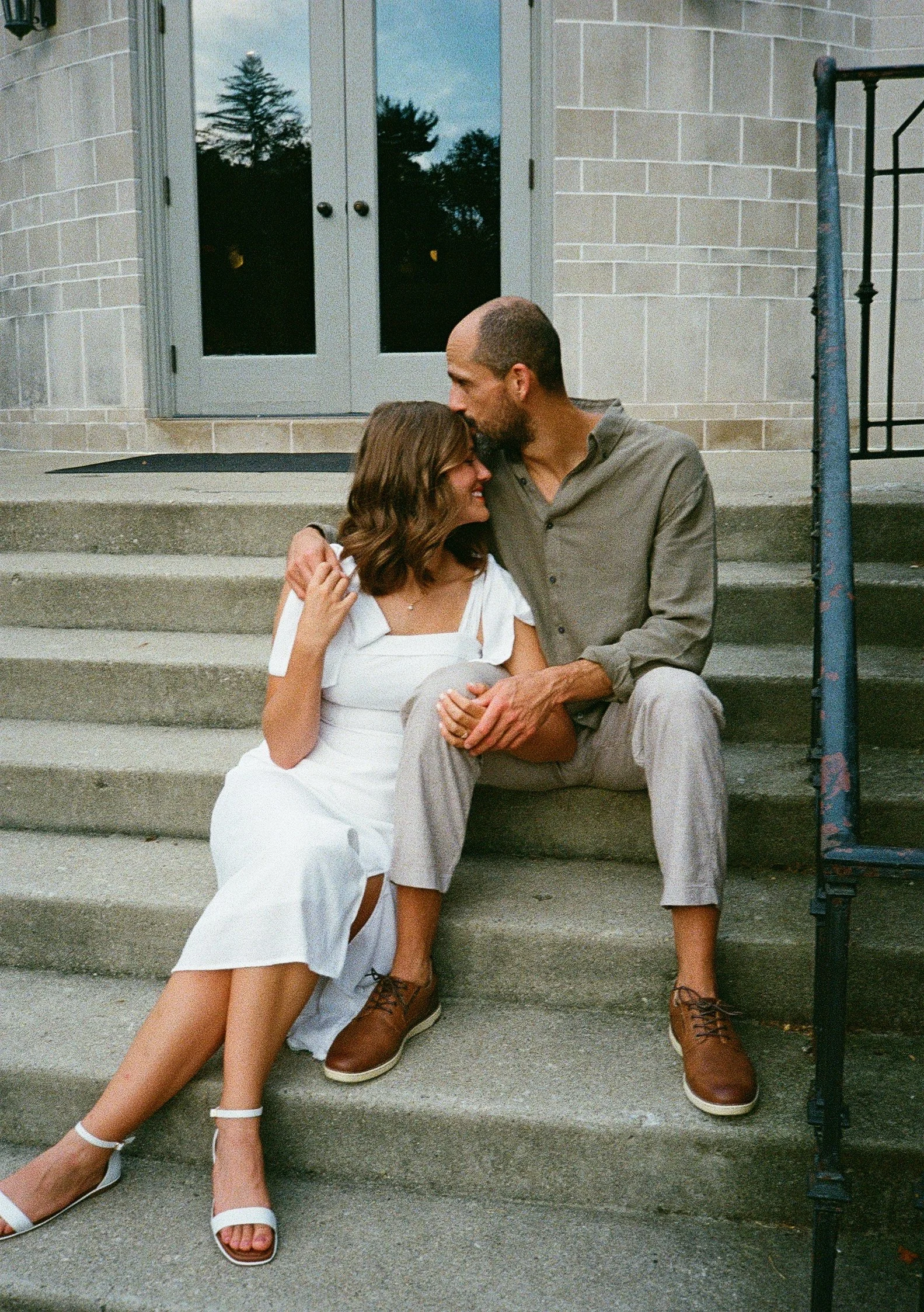 A couple sitting on steps outside a building. The woman is wearing a white dress and white heels, while the man is in a gray outfit and brown shoes. They are smiling and leaning close to each other. Columbus, Ohio engagement session.