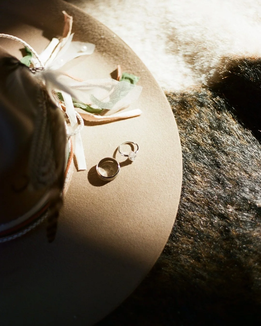 A pair of rings, a bouquet with ribbon and fabric, and a small bag on an oval beige surface, with a textured dark background.