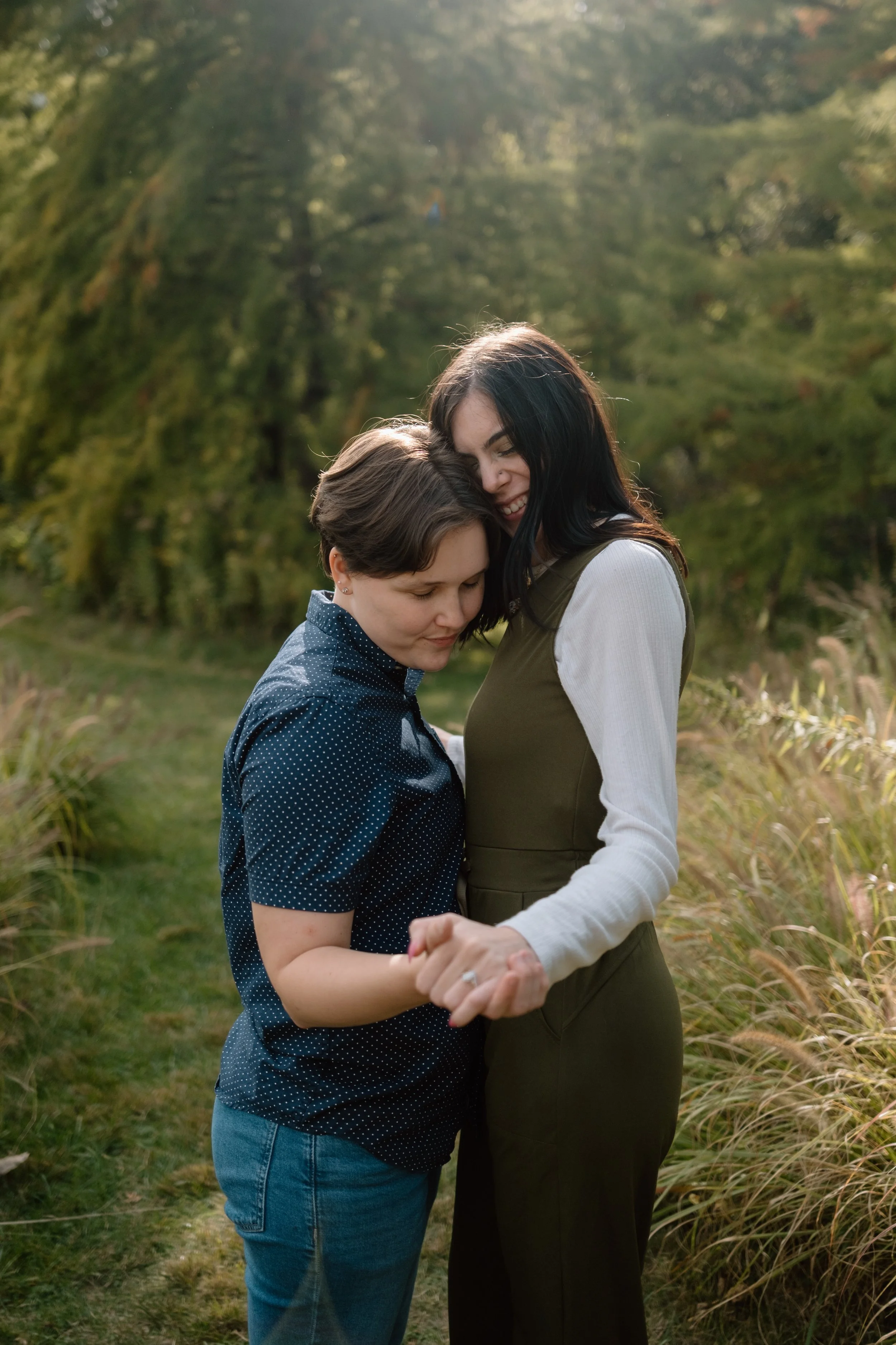 Two women sharing an intimate moment outdoors, standing close together on a grassy path surrounded by trees and greenery, holding hands and hugging.