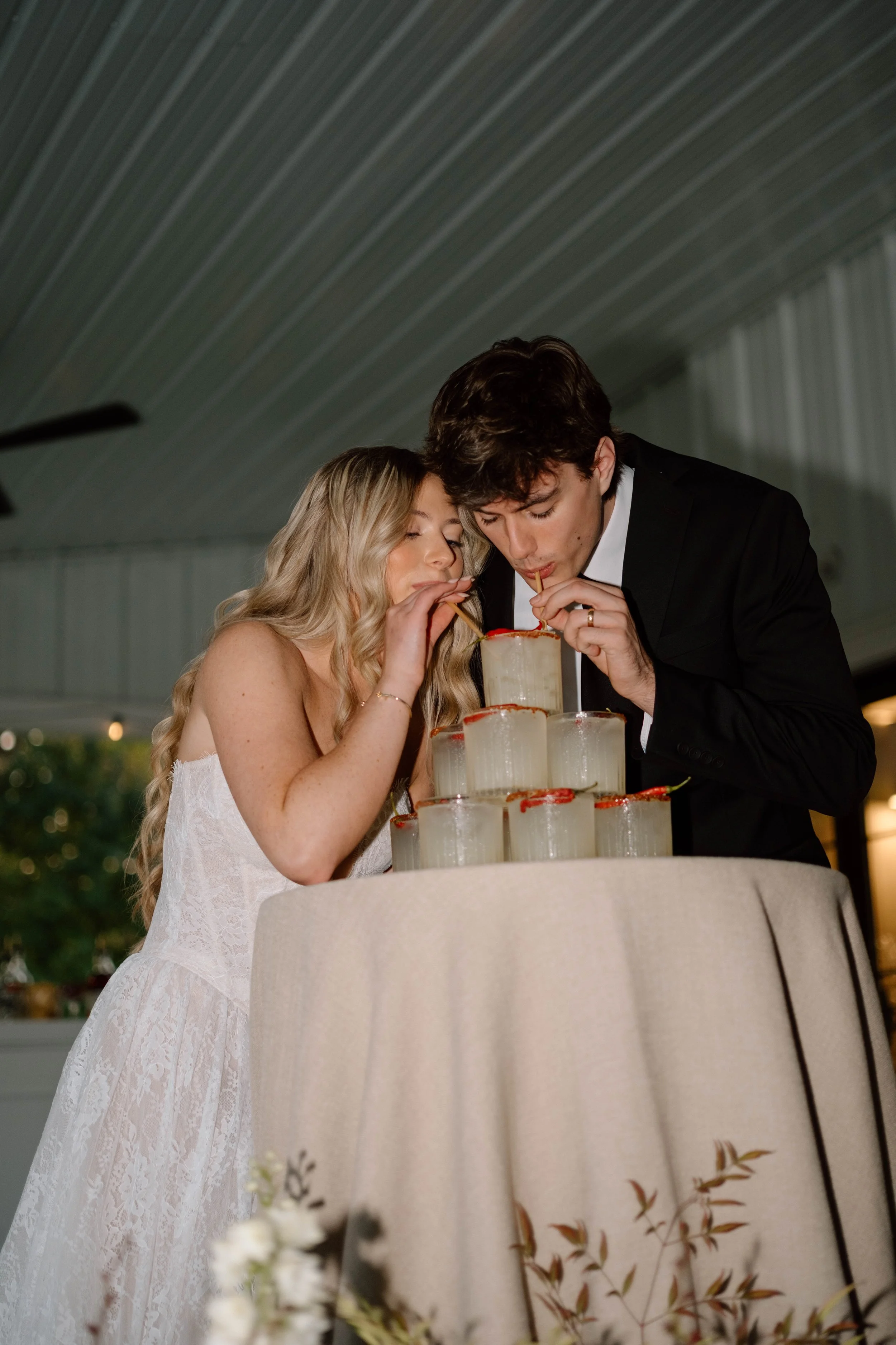 A bride and groom in wedding attire sharing a drink from a pyramid of glass containers during their wedding celebration.