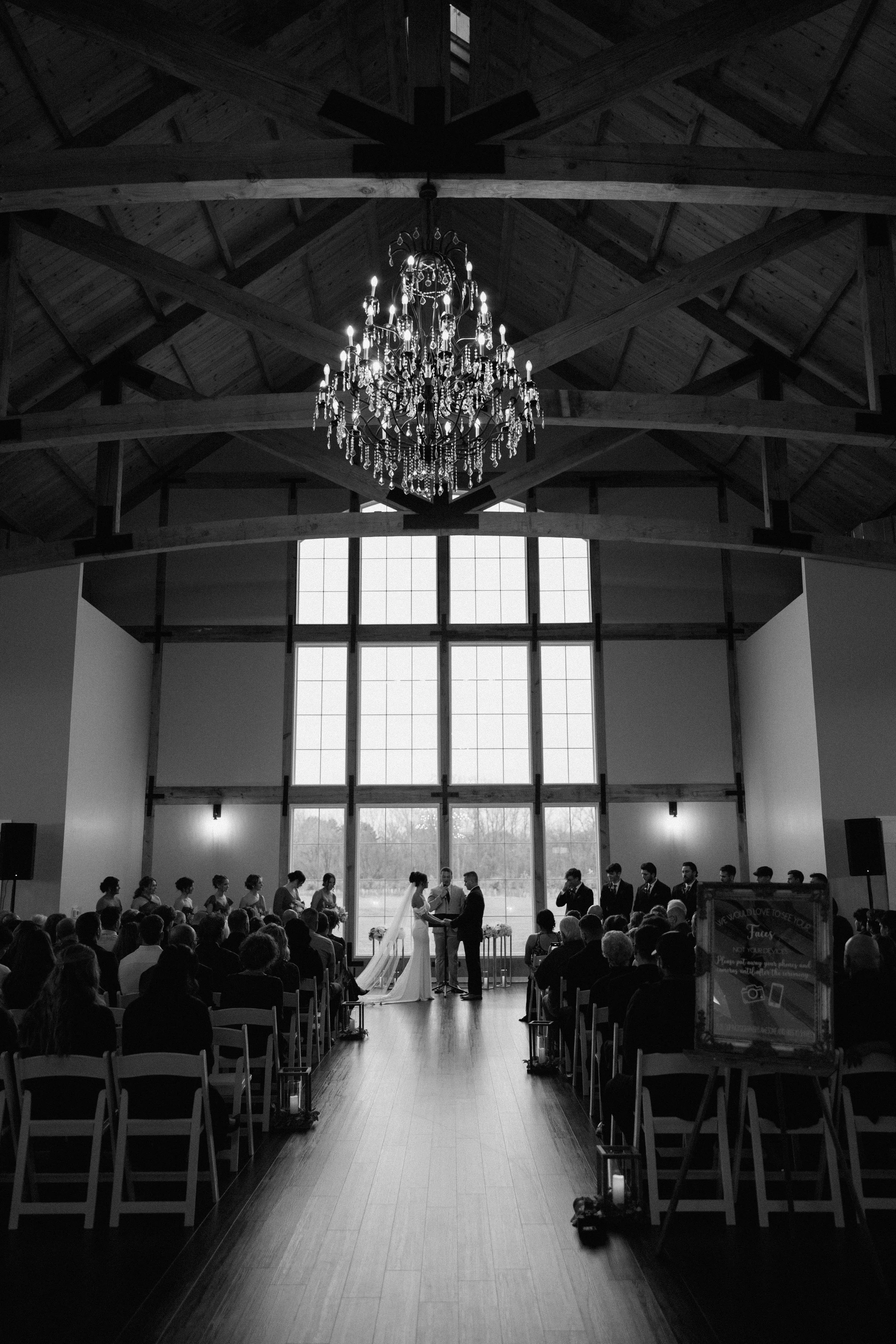 Black and white photo of a wedding ceremony inside a large, wood-beamed hall with a chandelier hanging from the ceiling. The bride and groom stand in front of an officiant, with bridesmaids and groomsmen on either side. Guests are seated on either si