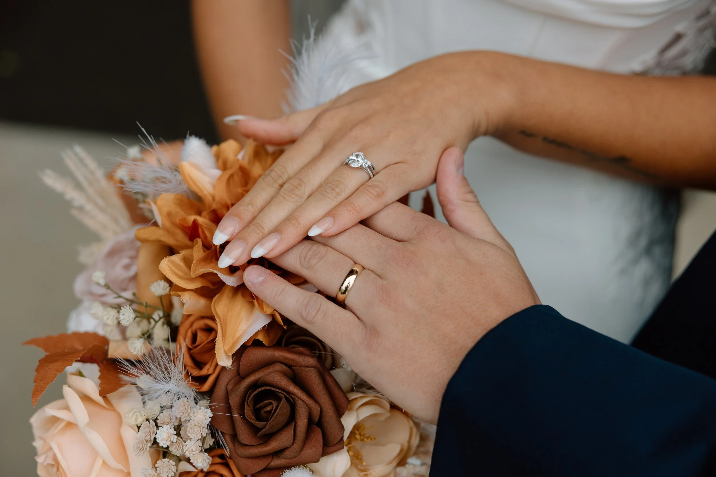 Close-up of a bride and groom's hands with wedding rings, holding a bouquet of orange, cream, and brown flowers, including roses and dried foliage.