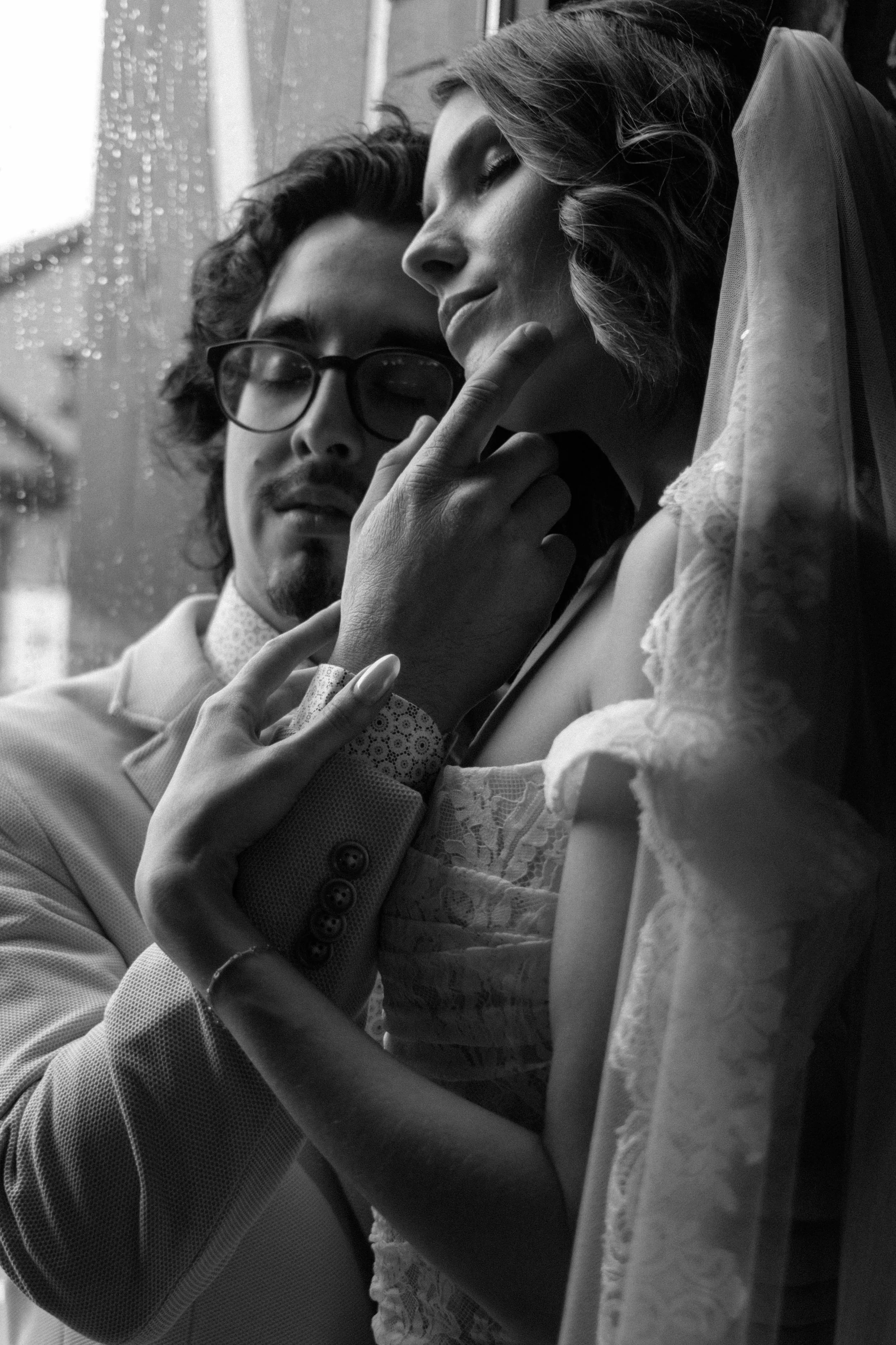 A black and white photo of a man and woman in a close, intimate moment. The man, wearing glasses and a light-colored suit, gently touches the woman's chin while she smiles softly, her eyes closed. She wears lace clothing and a veil.