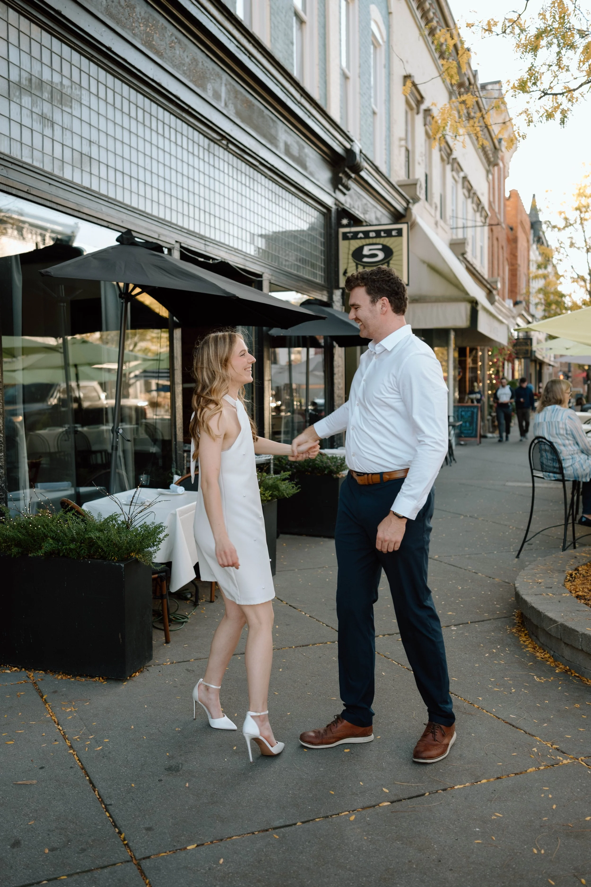A couple dancing on a city sidewalk outside a restaurant, holding hands and smiling at each other, with other people sitting at outdoor tables in the background. Northville, Michigan engagement session.