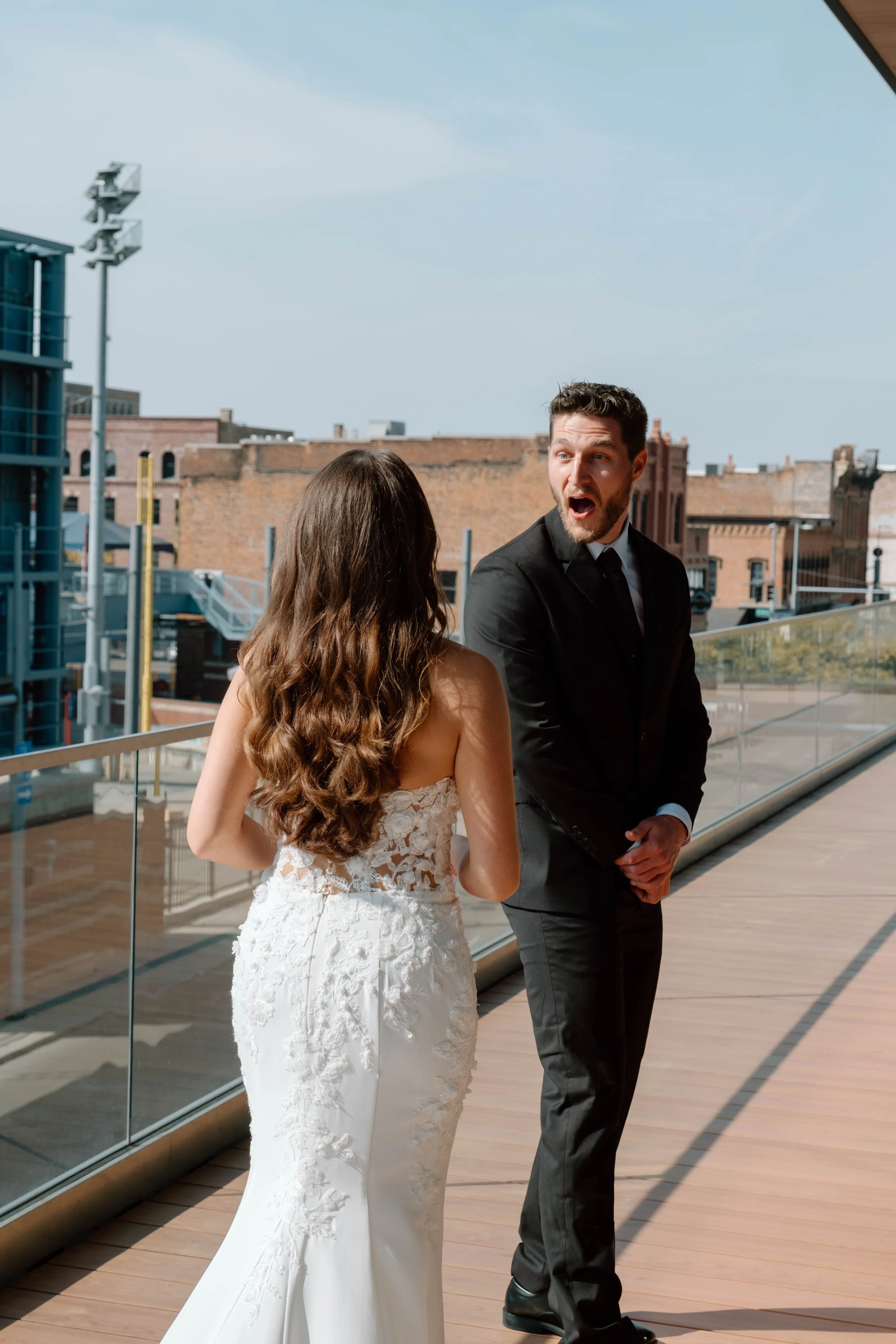 A man in a black suit reacting with surprise or excitement to a woman in a white wedding dress on a rooftop deck with city buildings in the background.