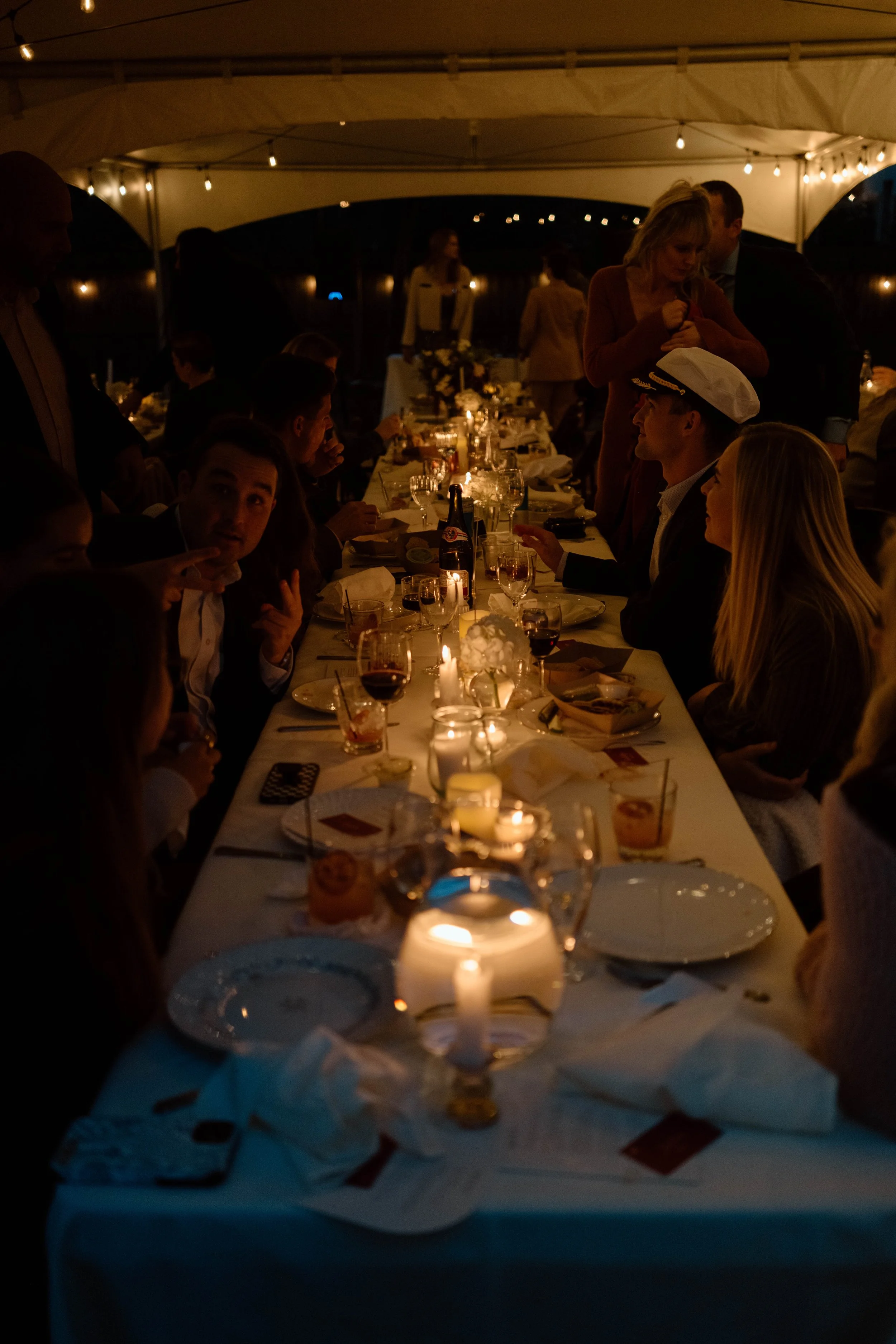 Nighttime dinner party under a tent with a long table decorated with candles, flowers, and various drinks, with guests dressed formally and engaging in conversation. Columbus, Ohio backyard wedding.