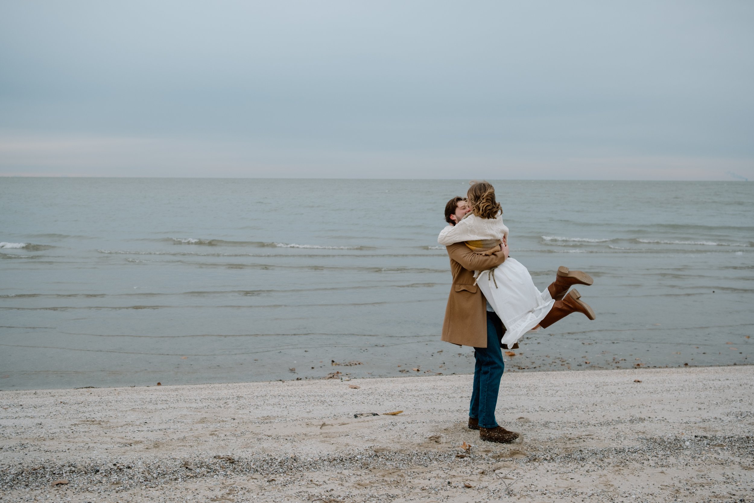 A man in a brown coat lifting a woman in a white dress on the beach, with the ocean and cloudy sky in the background.