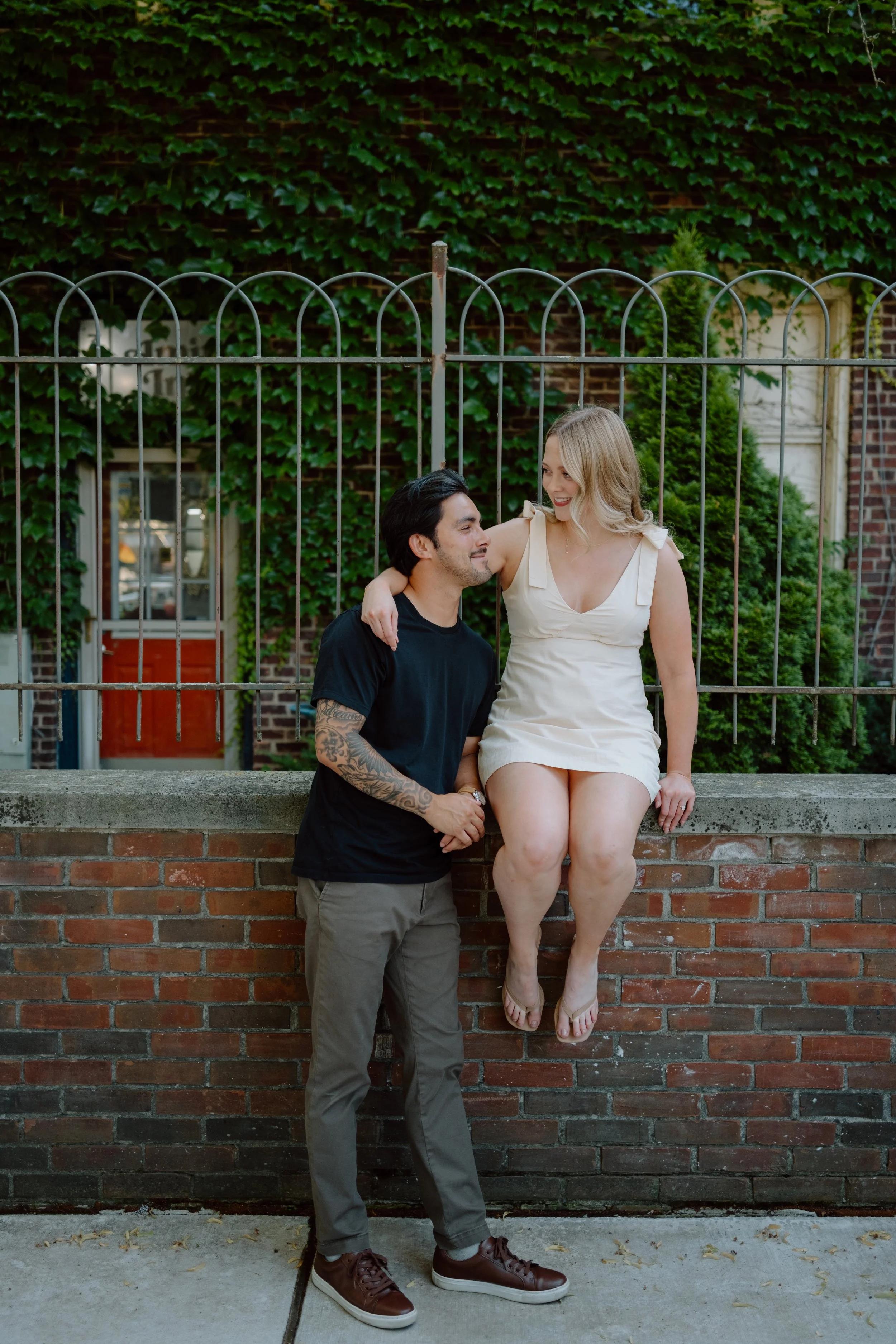 A man and a woman sharing a moment outdoors in front of a brick wall with green ivy. The woman is sitting on the wall, smiling at the man who is standing beside her, looking up at her. The woman has blonde hair, is wearing a white dress, and is baref
