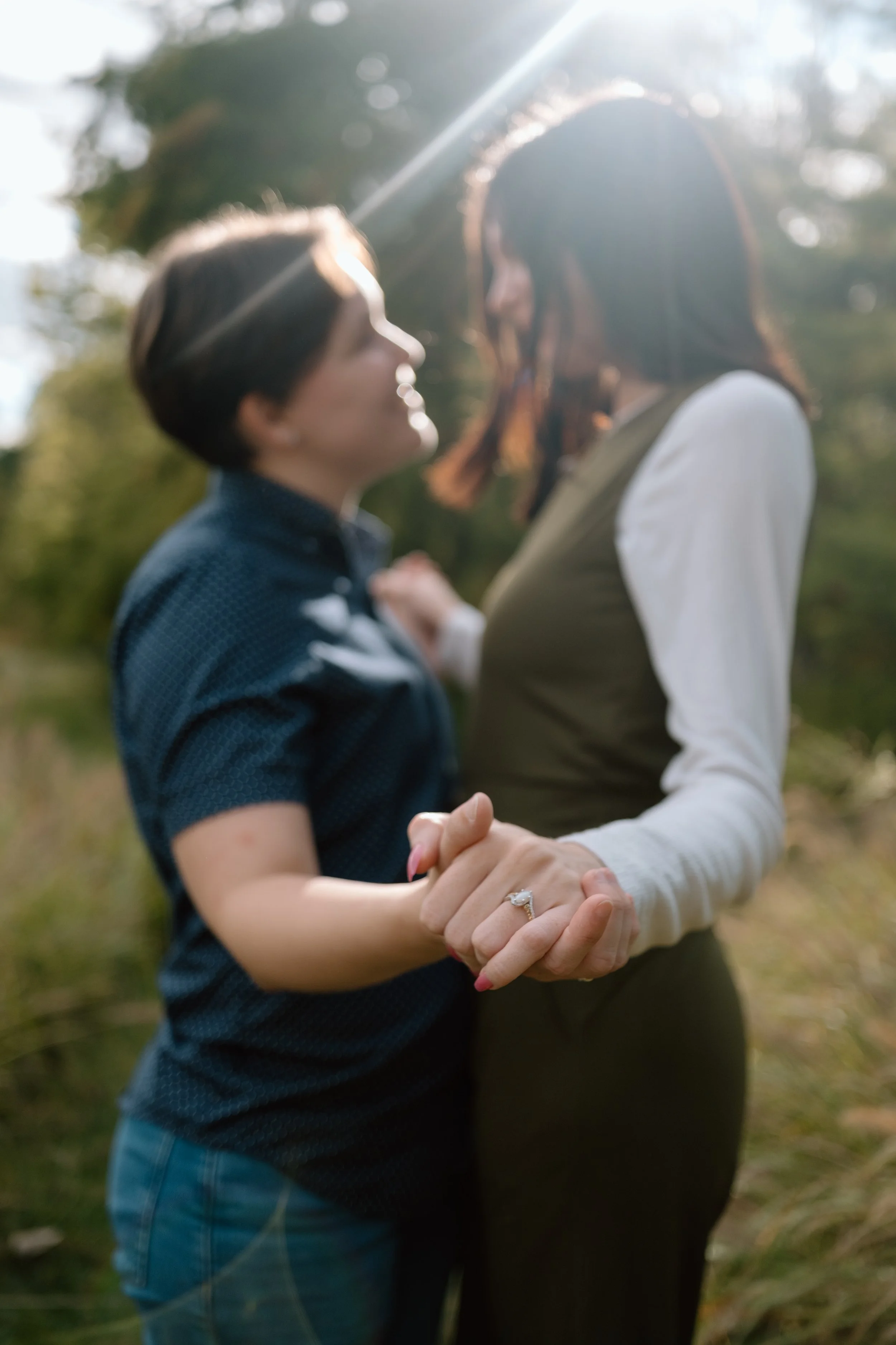 Two women are holding hands and facing each other in an outdoor setting, with one woman wearing a dark blue shirt and the other a white and olive top. They are smiling and looking into each other's eyes, with natural sunlight shining behind them.
