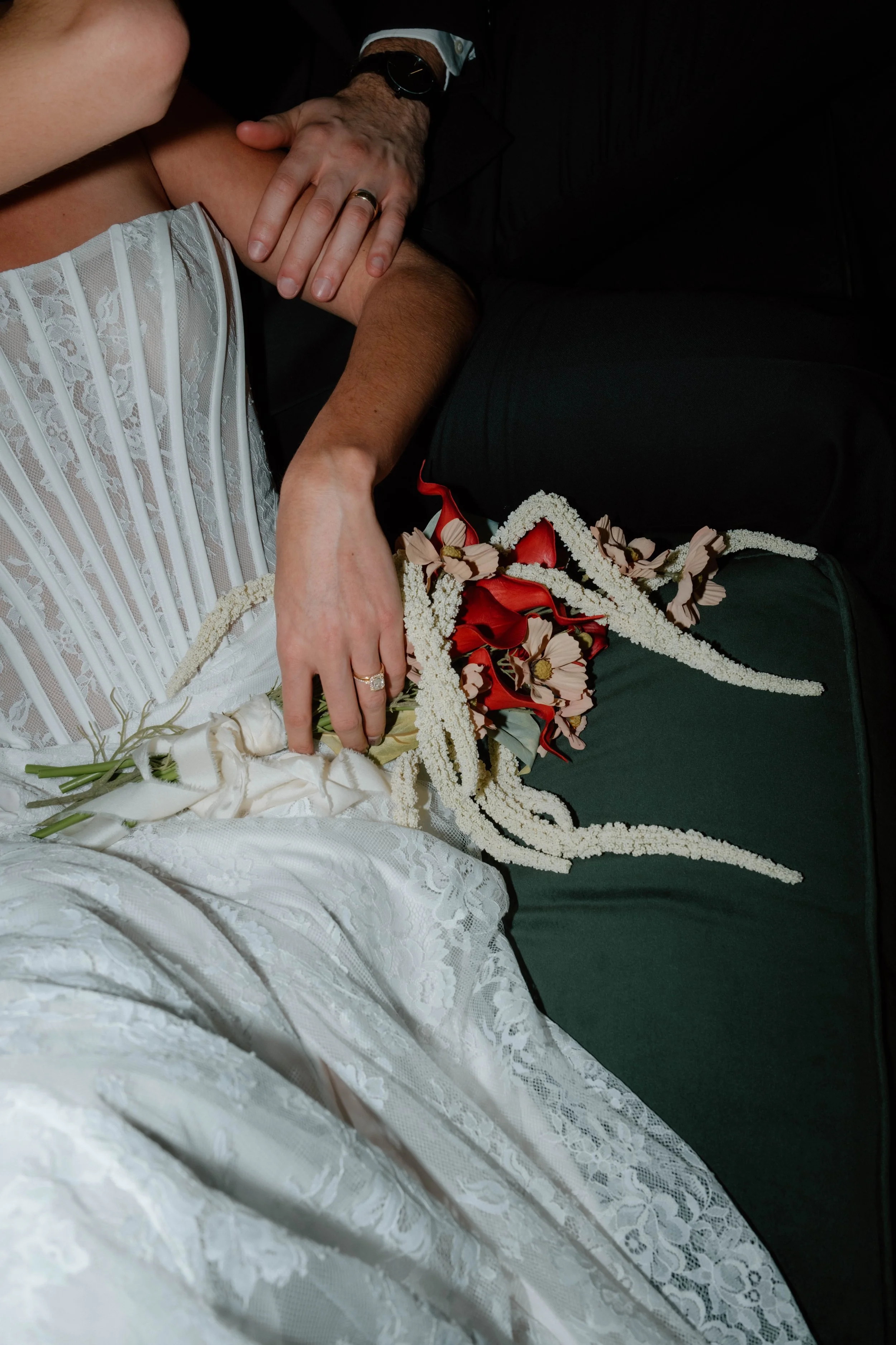 Close-up of a newlywed couple during their wedding, with the bride laying in a white wedding dress and the groom in dark formal attire. The groom's hand rests on the bride's shoulder, and a bouquet with red and white flowers is placed on her lap.