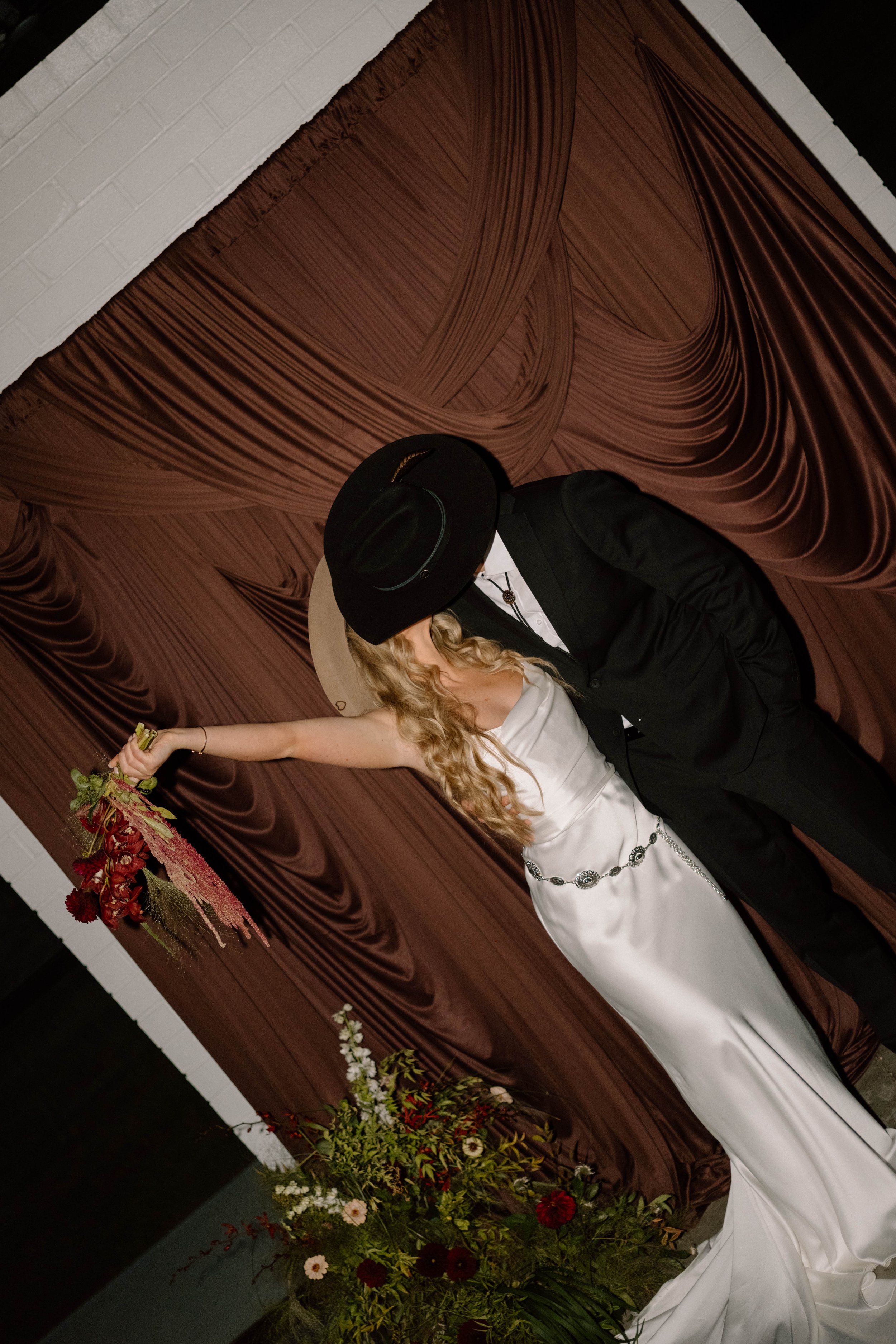 A couple dressed in formal wedding attire, with a woman in a white gown holding a bouquet and a man in a black tuxedo with a hat, standing in front of brown draped fabric and floral arrangements.