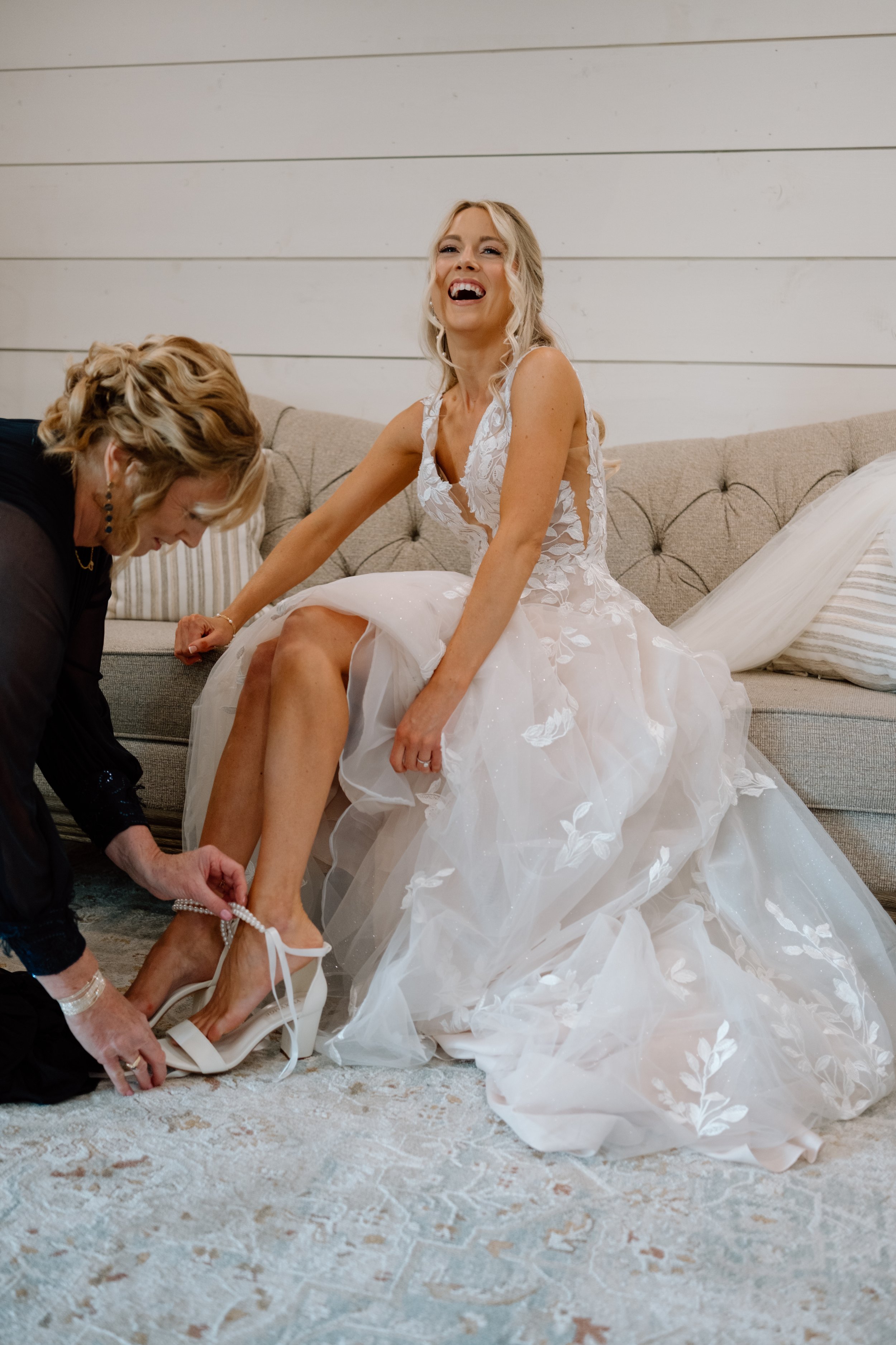 Bridal bride laughing while sitting on a couch, having her white wedding shoes put on by an older woman.