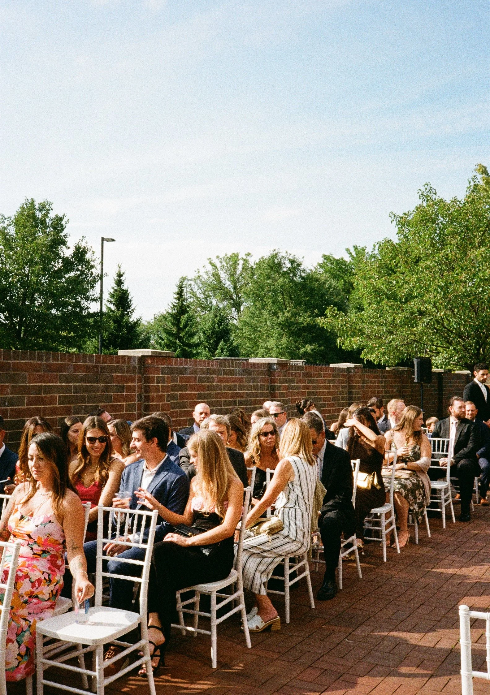 People dressed in formal attire sitting on white chairs outdoors, with a brick wall and green trees in the background, at a wedding on a sunny day in Chicago, Illinois.