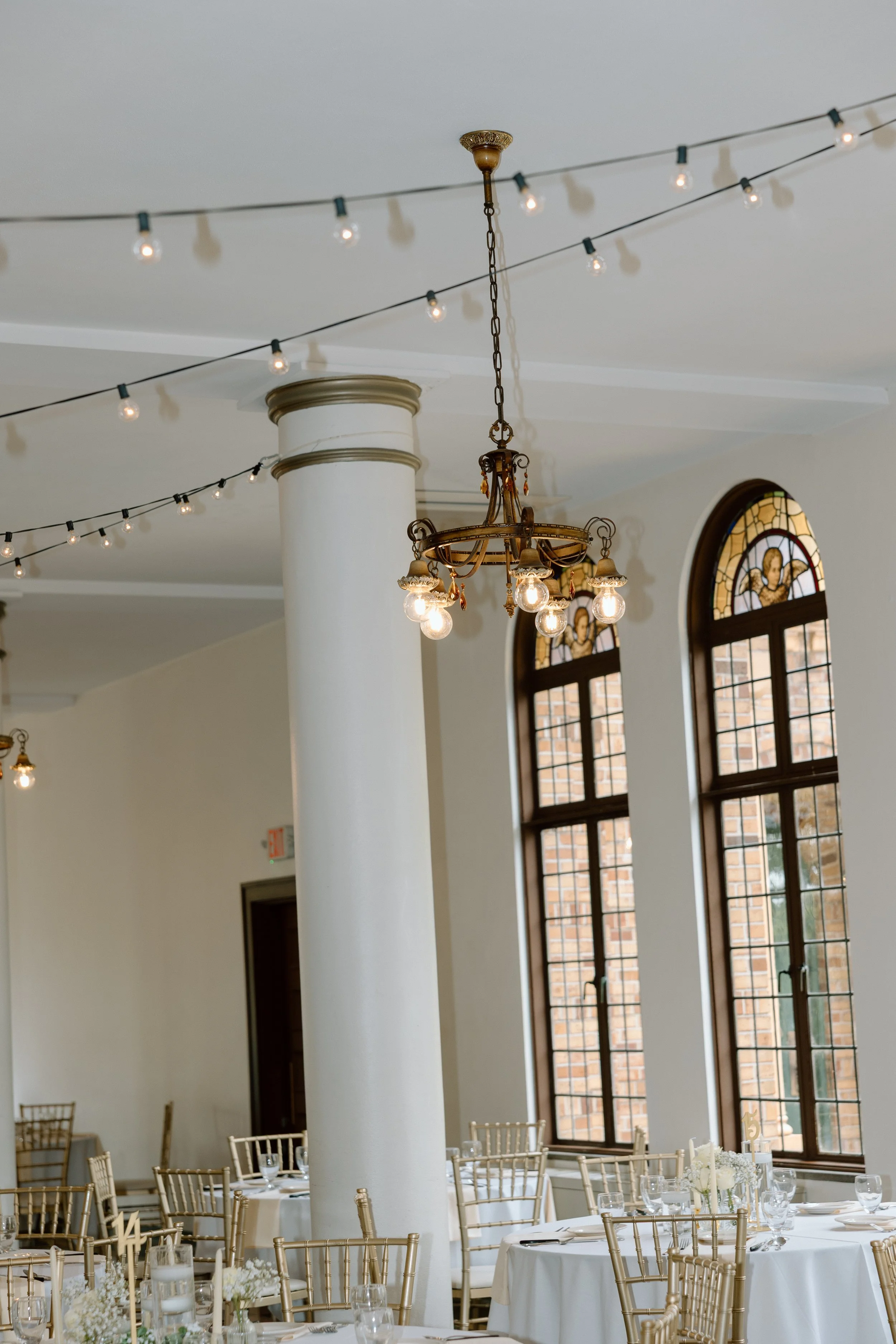 Elegant event hall with round tables, gold chairs, large arched windows with stained glass, and hanging string lights and chandeliers.