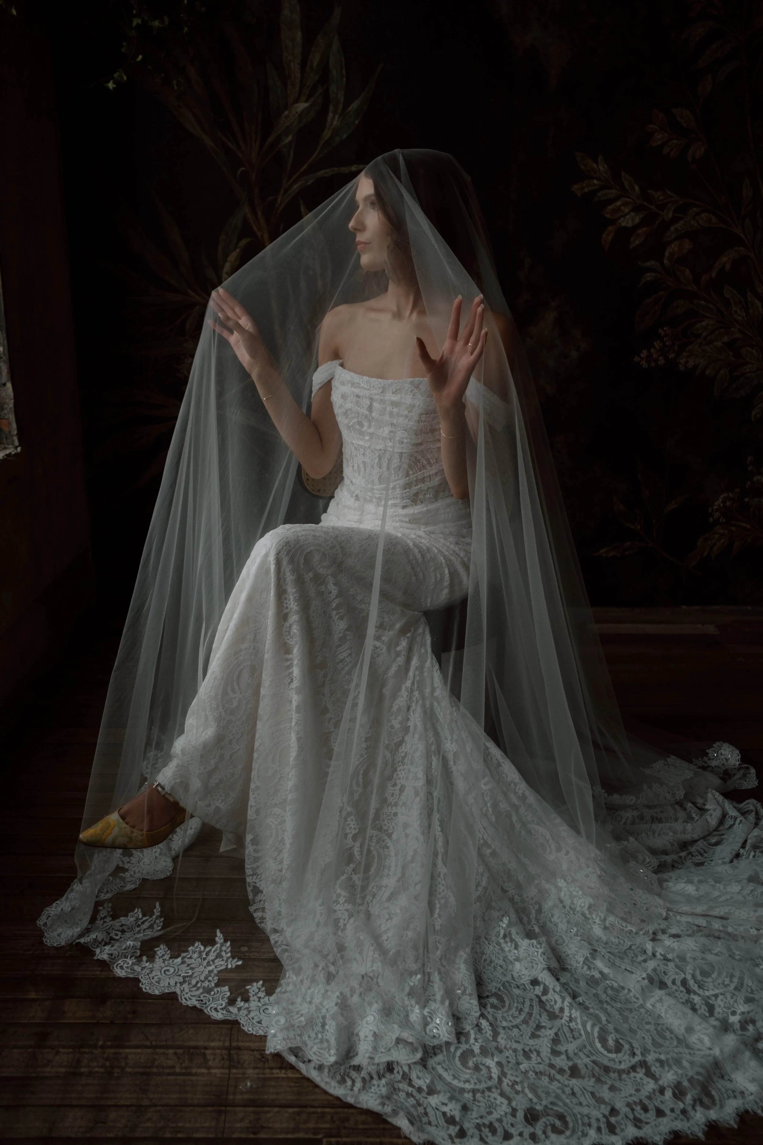 A woman in a lace wedding dress sitting behind a sheer veil, posed against a dark floral background.