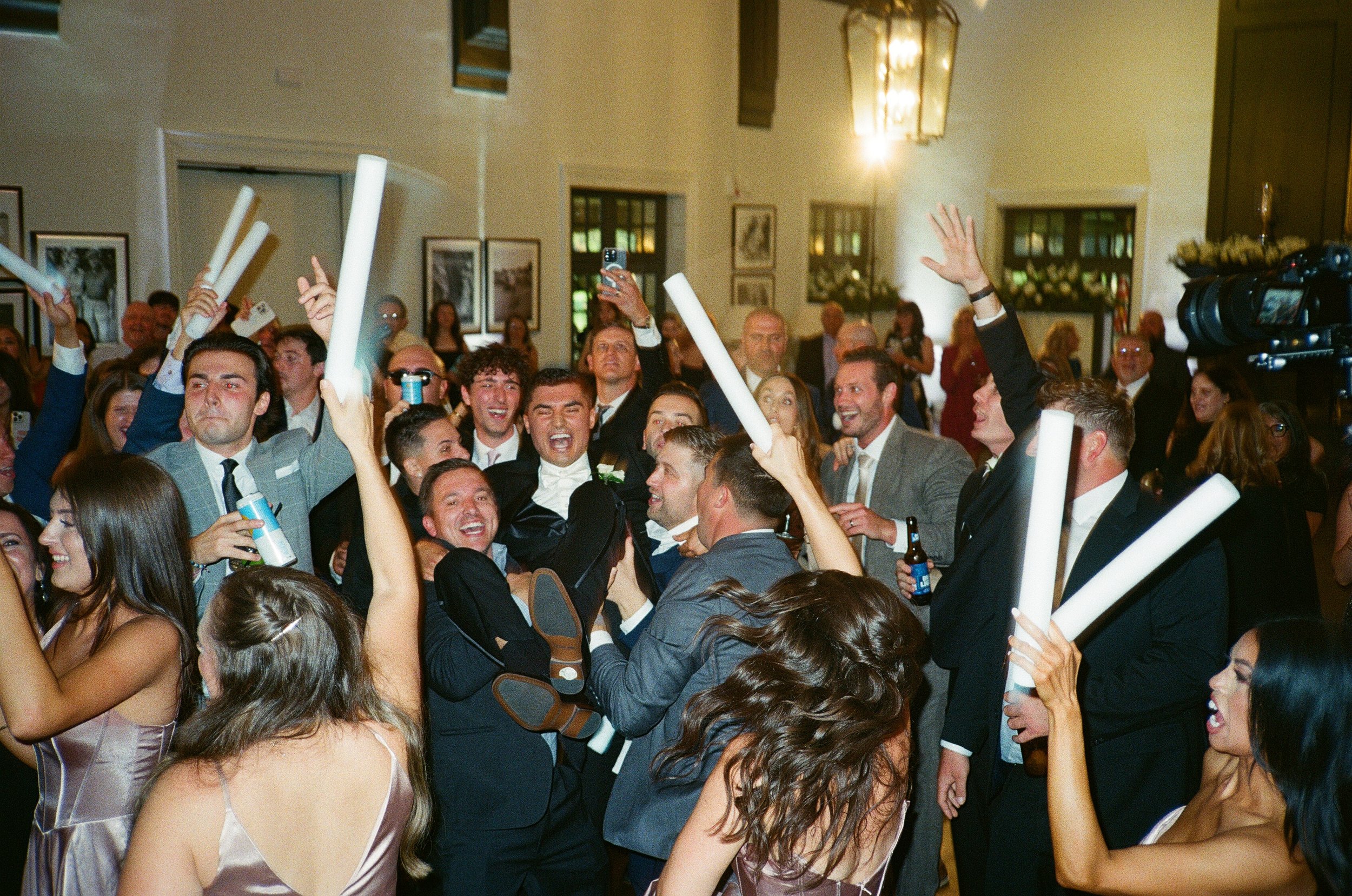 Group of people celebrating at a wedding reception, some holding glow sticks, dancing and smiling, with a bride in a white dress and groom in a tuxedo at the center.
