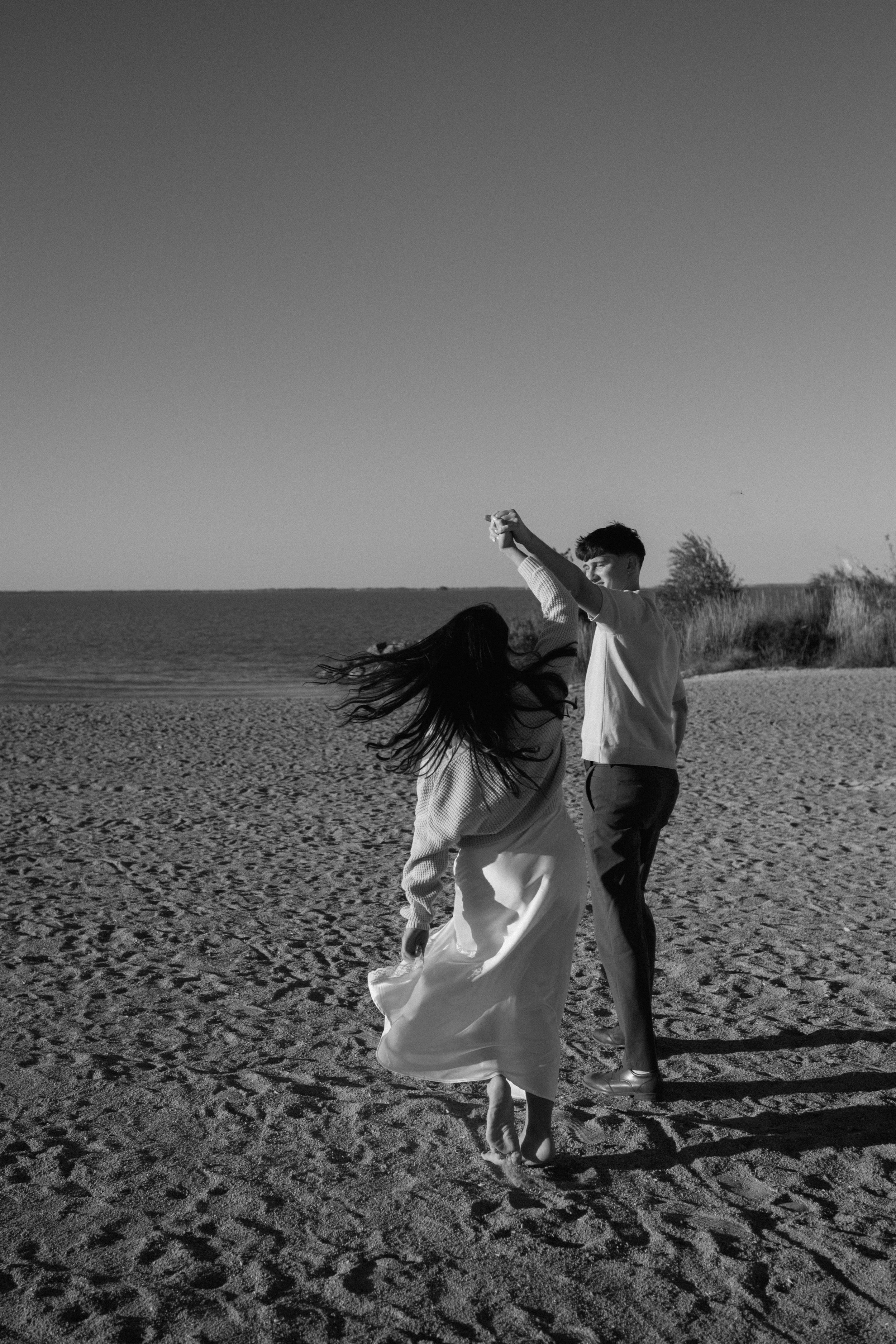 A black-and-white photo of a man and woman dancing on a sandy beach, holding hands, with the woman spinning and hair flowing.