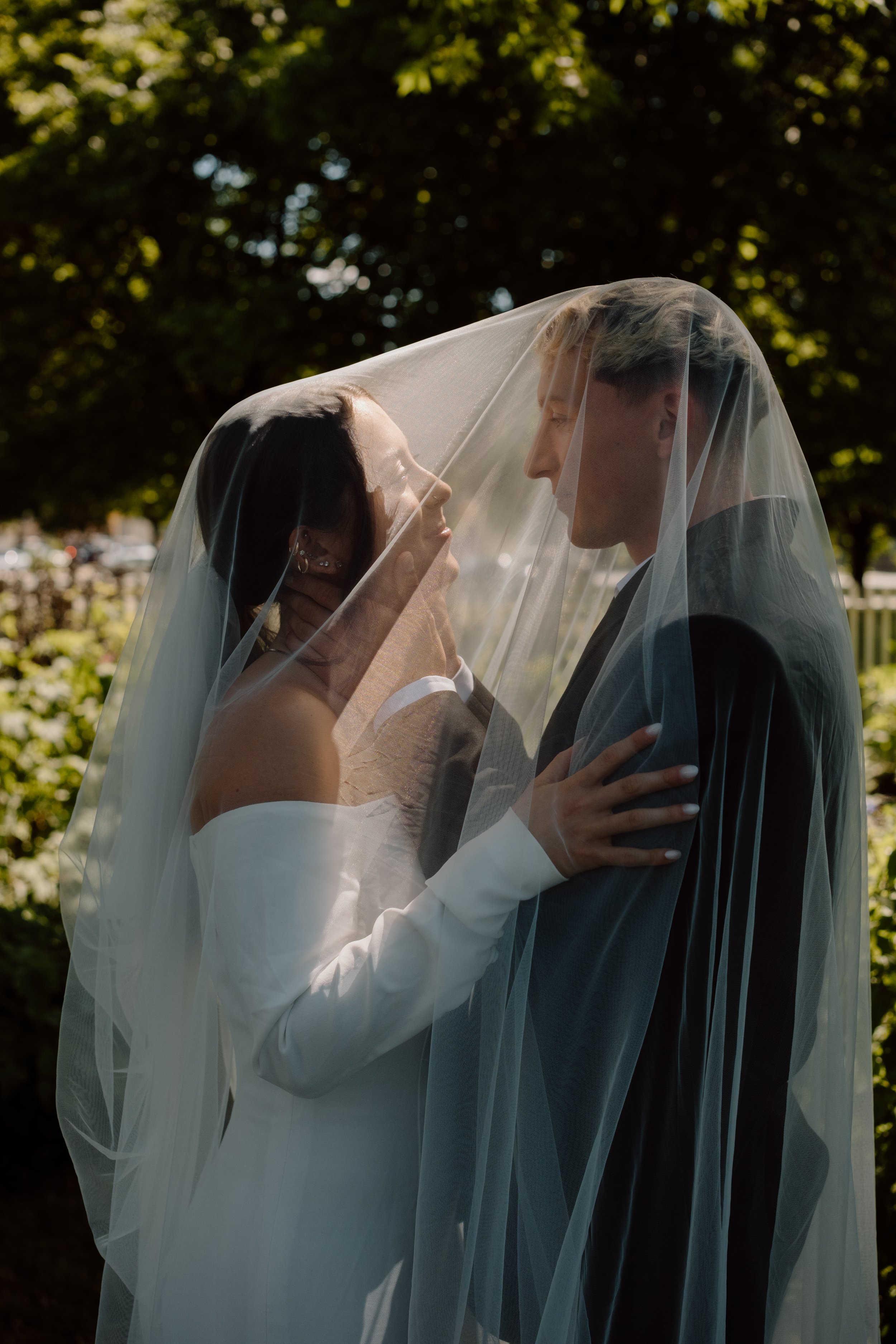 Bride and groom standing close under a veil outdoors, gazing at each other affectionately.