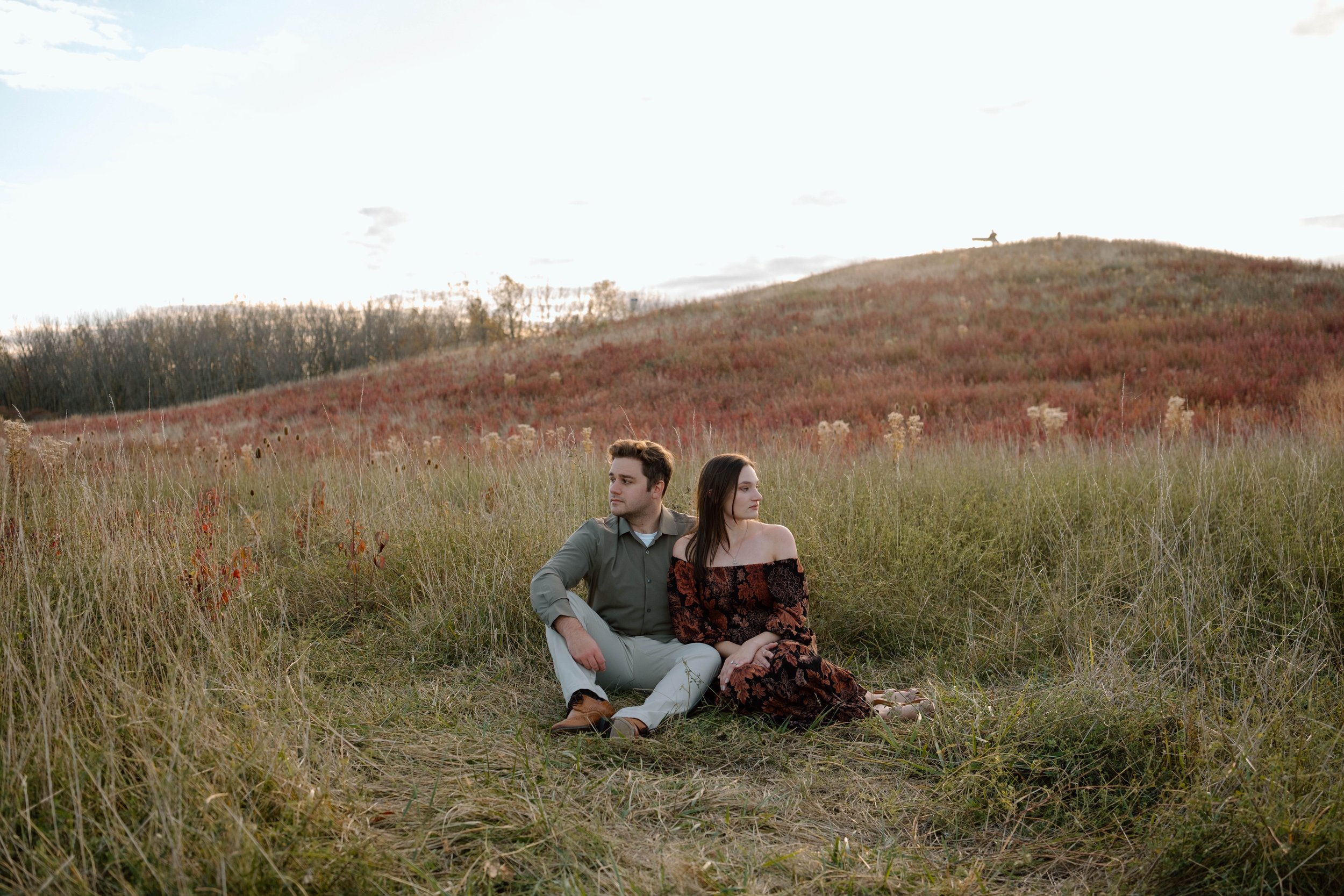 A young man and woman sitting together in a field of tall grass, with a hill covered in reddish foliage and a small dog statue on top in the background