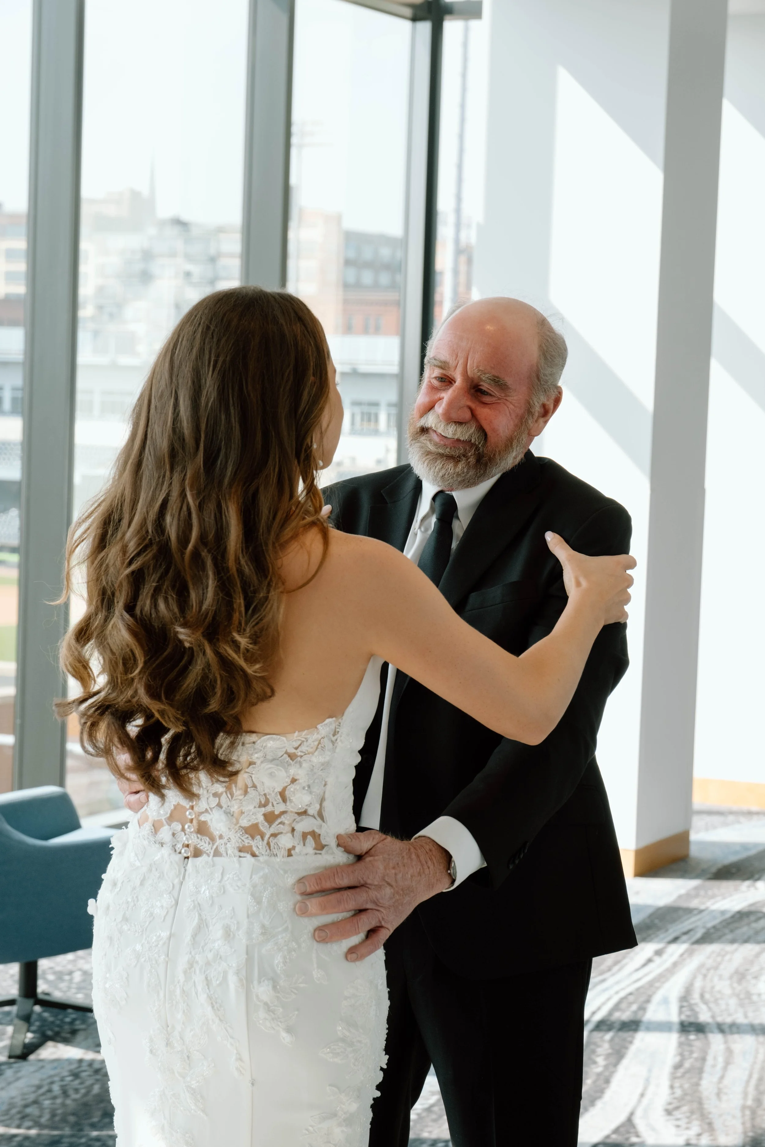 A bride and an older man, likely her father, share a dance in a bright, modern room with large windows.